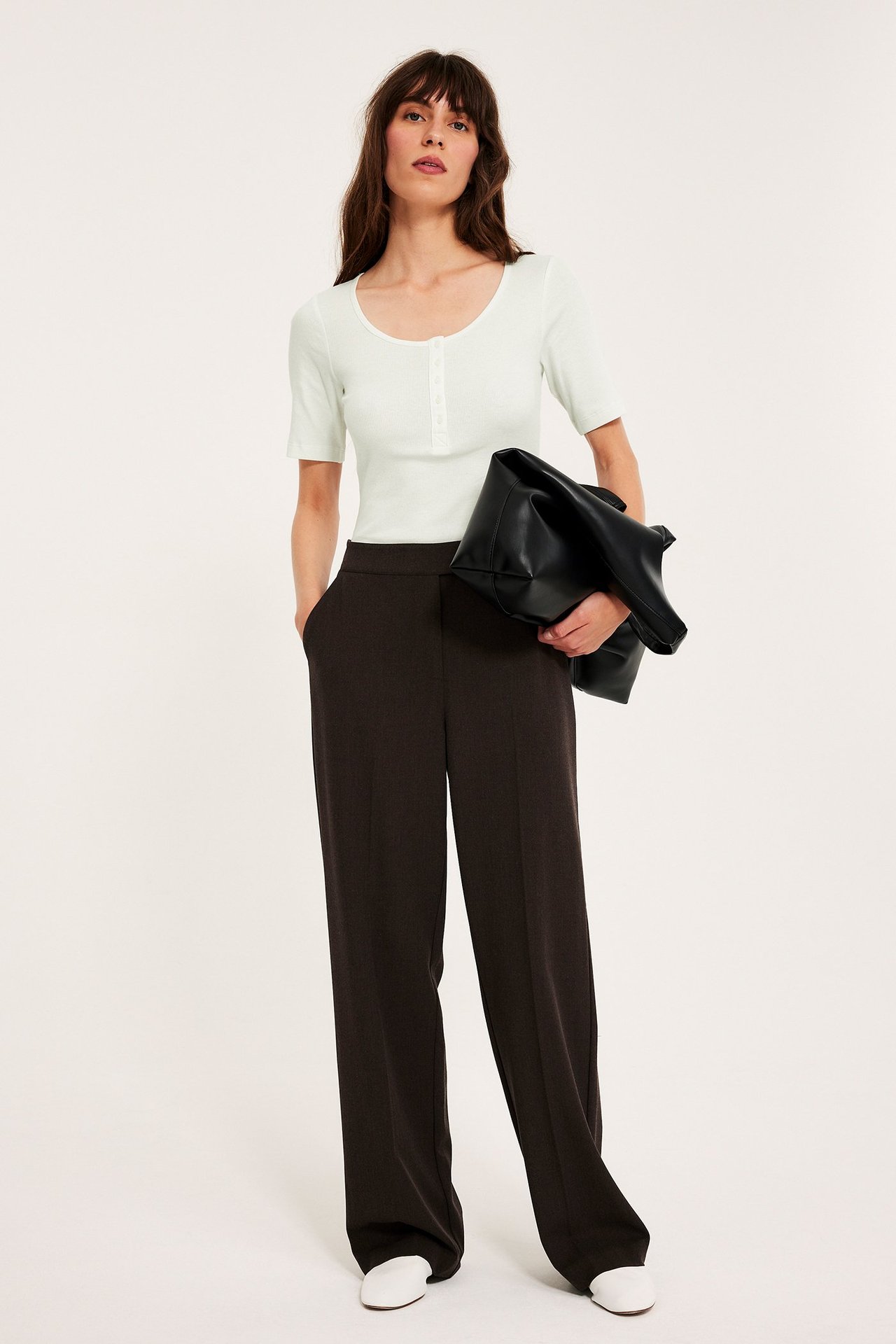 Edestä: Nainen luonnonvalkoisessa ribatussa lyhythihaisessa topissa napeilla, tummanruskeissa housuissa, valkoisissa kengissä, pitäen mustaa laukkua.