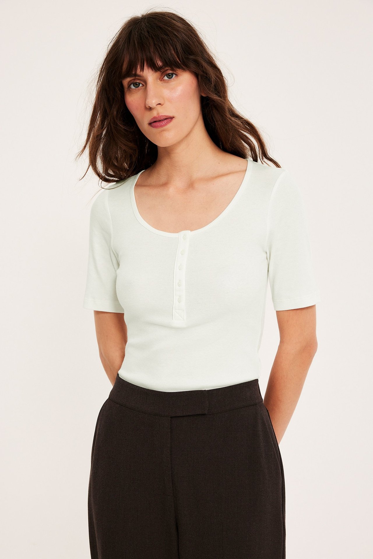 Kobieta w dopasowanym, prążkowanym topie off-white z krótkim rękawem, guzikami i ciemnoszarych spodniach.