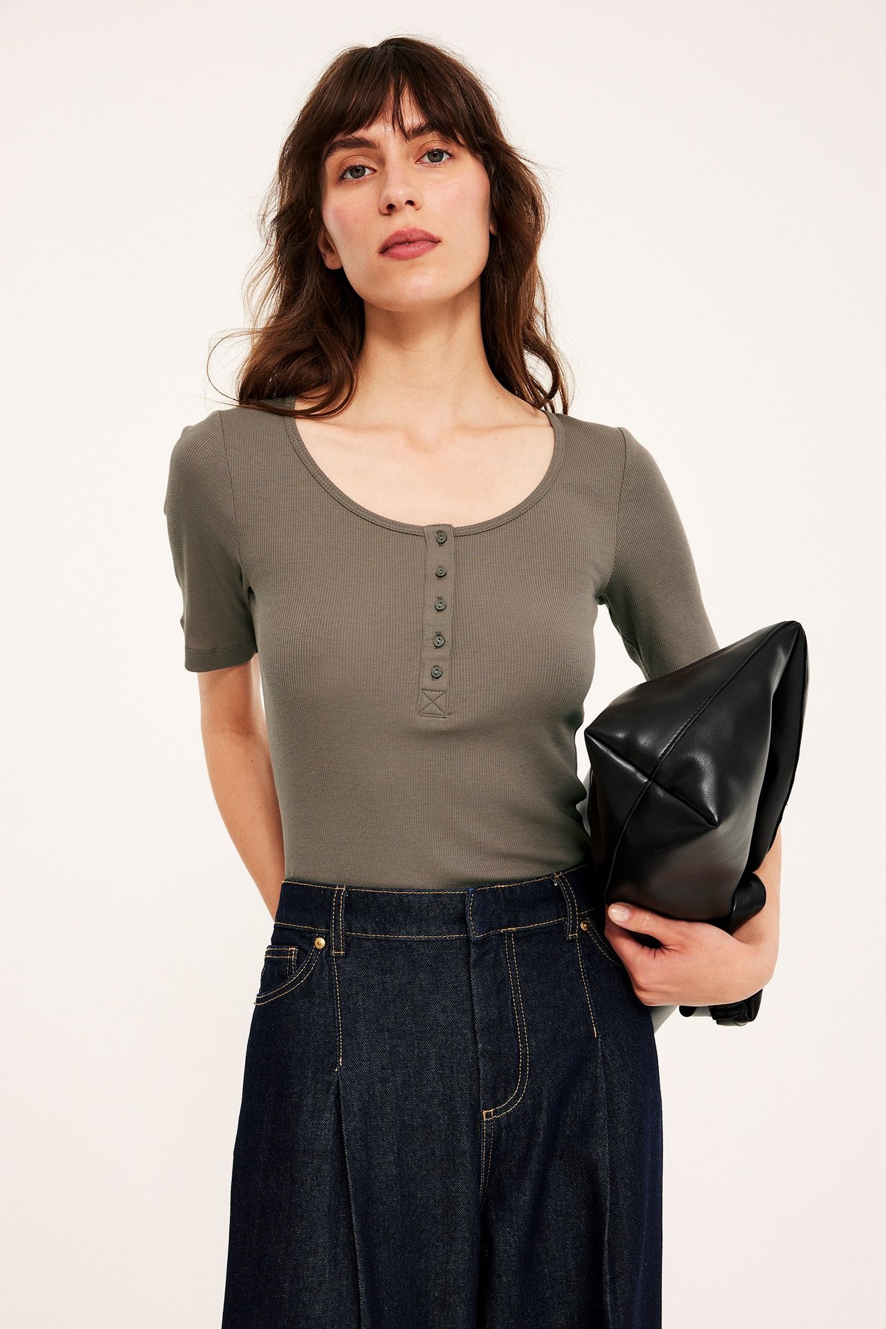 Widok z przodu: Kobieta w brązowym prążkowanym topie z krótkim rękawem, ciemnych dżinsach, trzyma czarną torebkę.