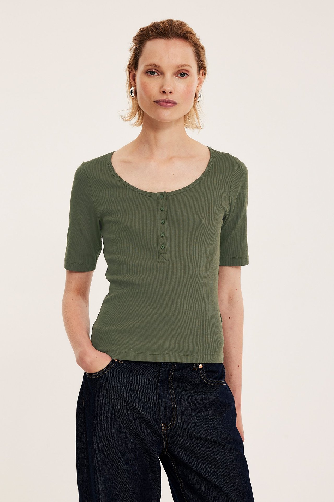 Widok z przodu: Kobieta w zielonym prążkowanym topie z guzikami, ciemnych dżinsach i srebrnych kolczykach.