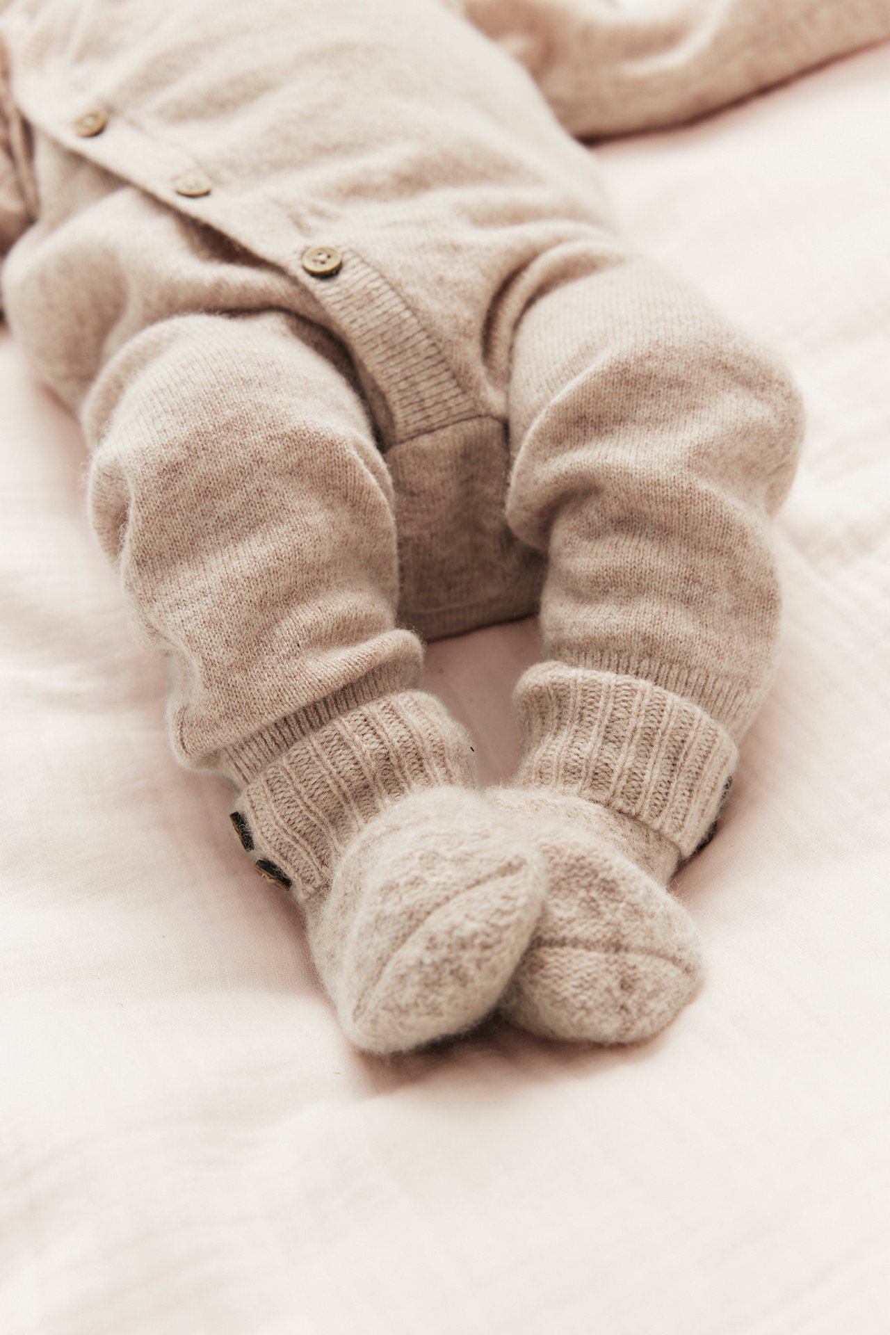 Nærbilde av en baby i en beige melert omslagsoverall i ull-kasjmir med knapper og ribbestrikkede mansjetter.