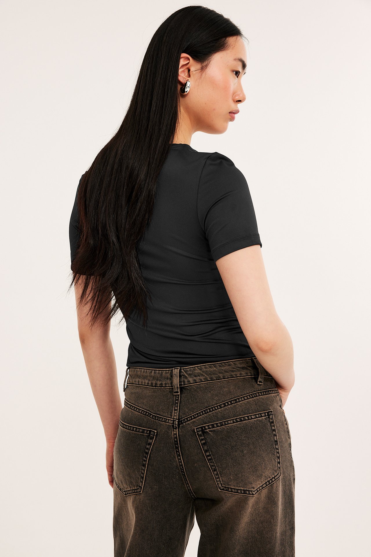 Bakifrån: kvinna i svart kortärmad t-shirt med rund hals, mörkbruna jeans och silverfärgade örhängen.