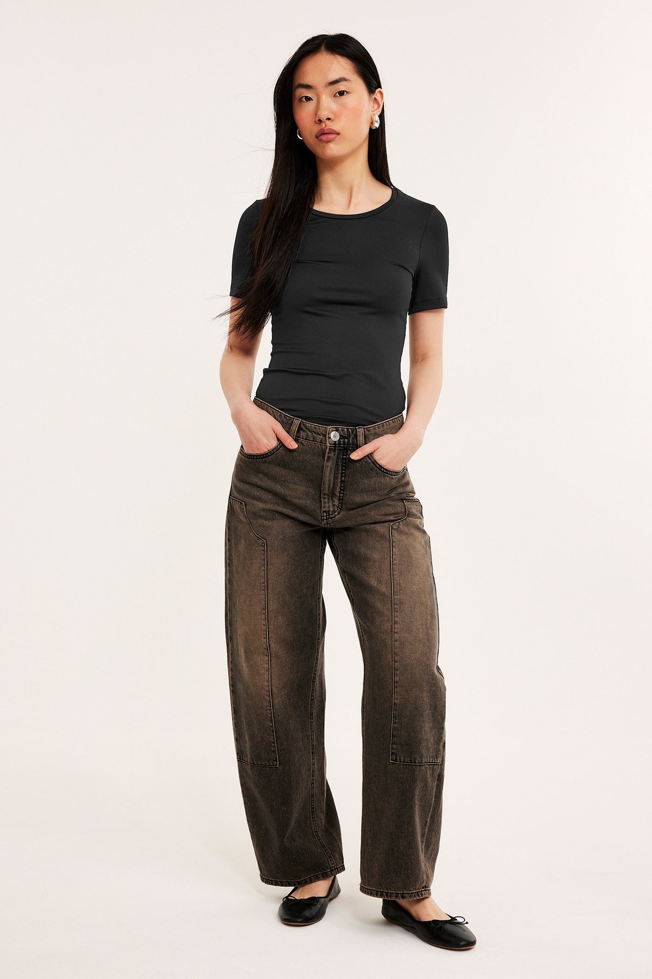 Frontbilde av kvinne i svart kortermet t-skjorte med rund hals, mørkebrune vide jeans og svarte ballerinasko.