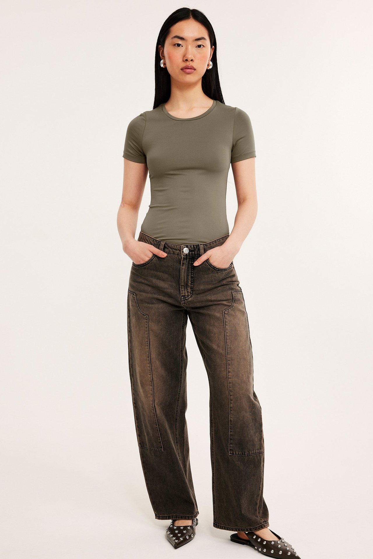 Frontbilde: kvinne i tettsittende khaki kortermet t-skjorte, mørkebrune vide jeans og svarte nagle-ballerinasko.