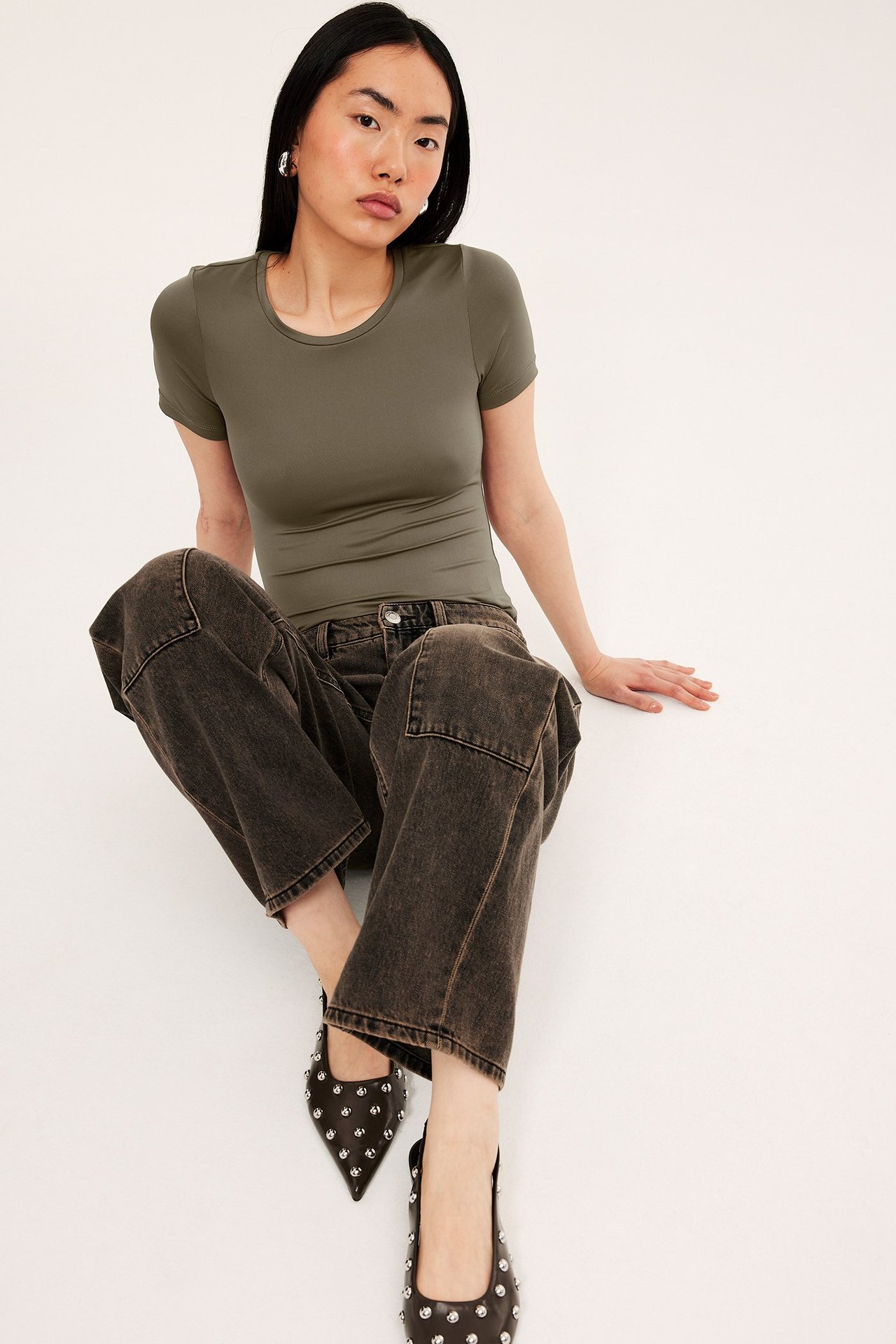 Forfra, kvinne i en figurnær olivengrønn kortermet t-skjorte, mørke jeans og svarte naglepyntede ballerinasko.