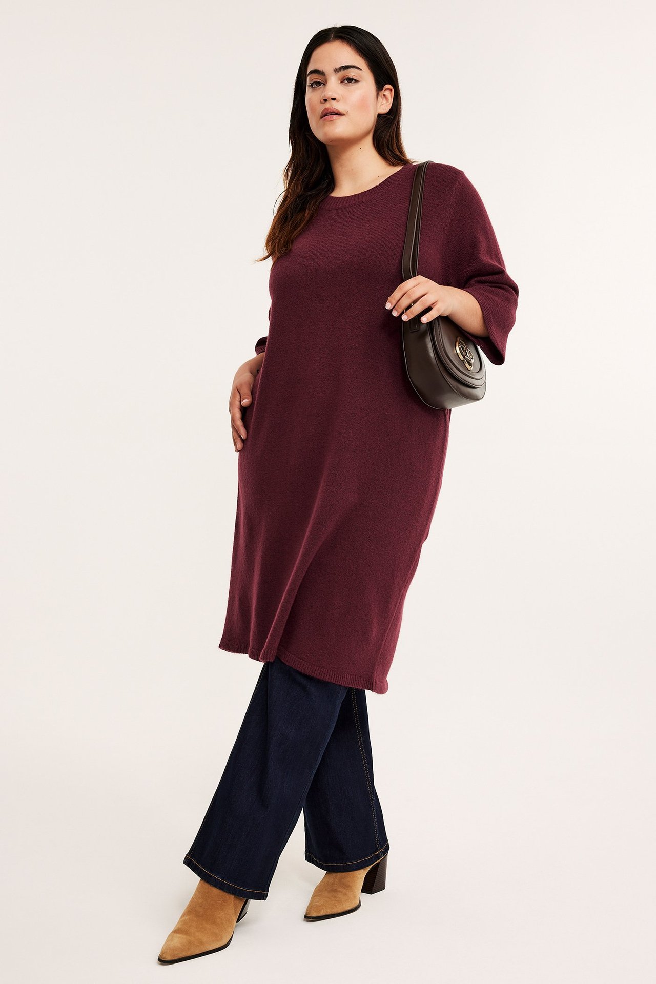 Frontvy av en kvinna i vinröd melerad stickad klänning, mörkblå jeans, beiga stövlar och brun väska.