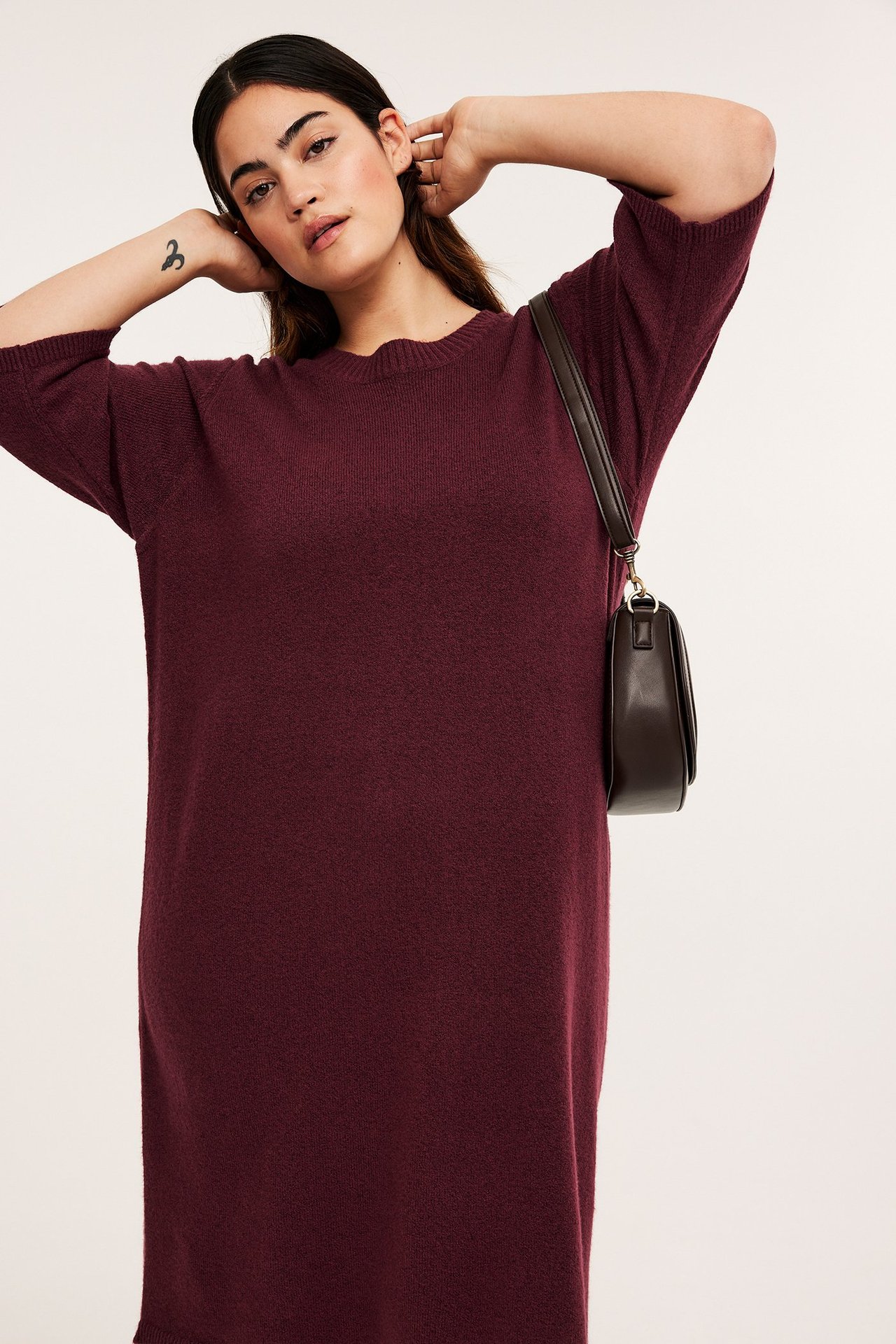 Frontvy av en kvinna i en vinröd melerad stickad klänning med 3/4-ärm och en mörkbrun axelväska.