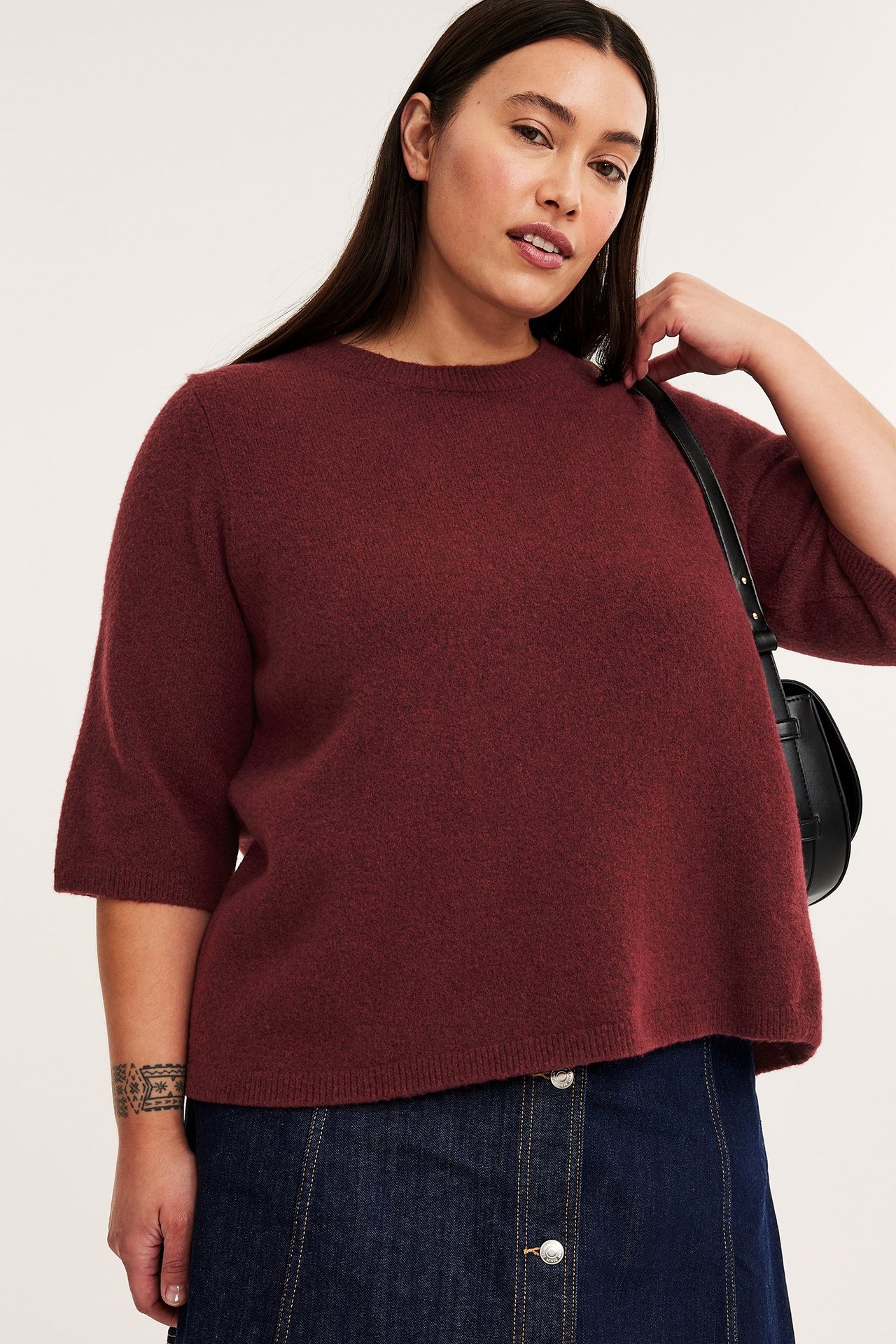 Etunäkymä: Nainen yllään meleerattu viininpunainen neuletoppi 3/4-hihoilla, tummansininen farkkuhame ja musta laukku.