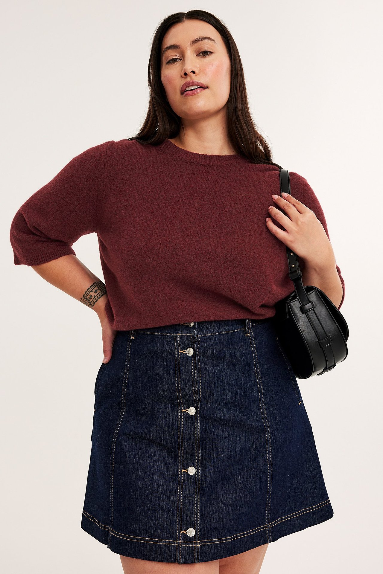 Edestä: Nainen viininpunaisessa melange neuletopissa, tummansinisessä farkkuhameessa ja mustassa käsilaukussa.