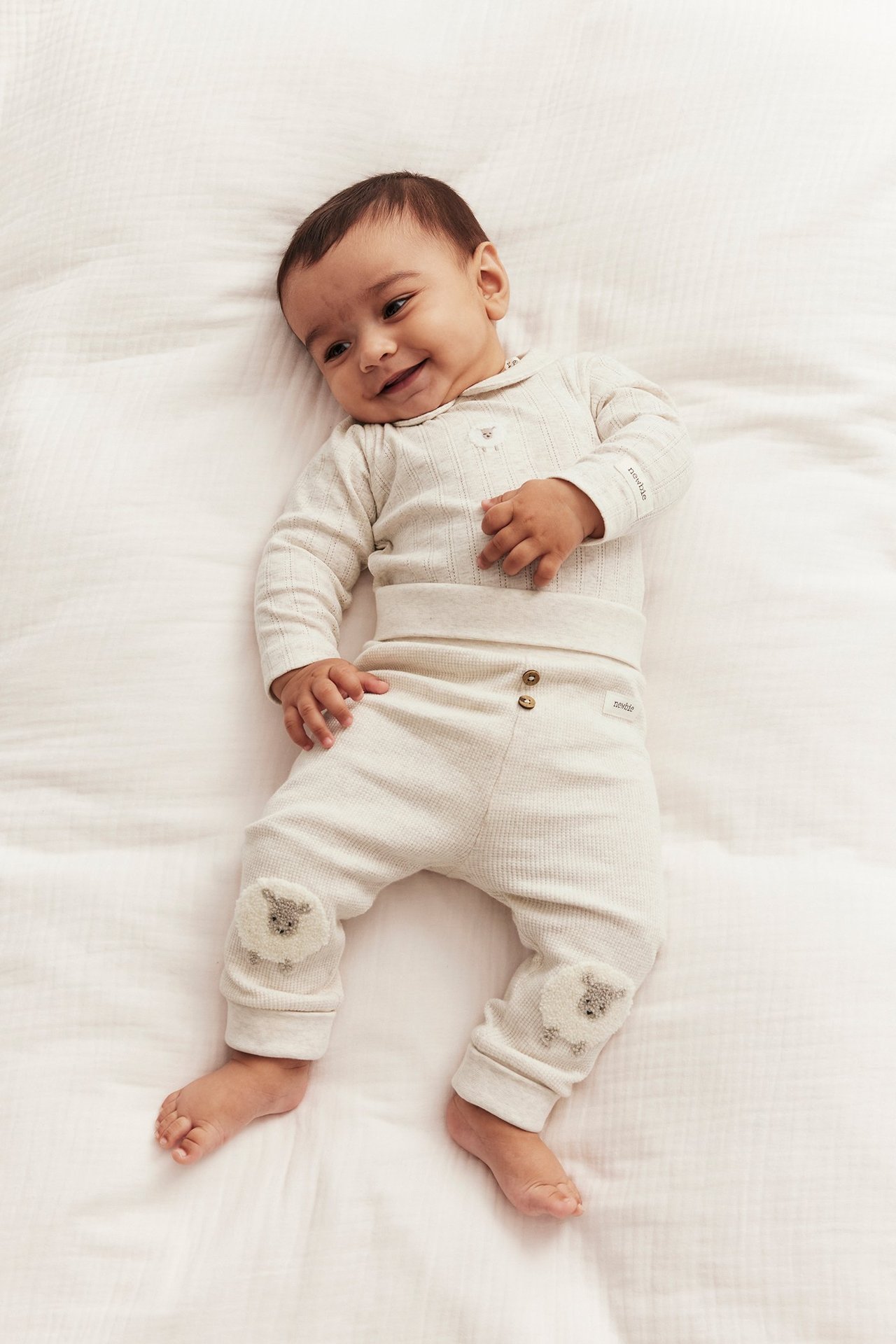 Baby i en beige melerad ribbad långärmad body med krage, bröstbrodyr och matchande byxor med får-applikationer.