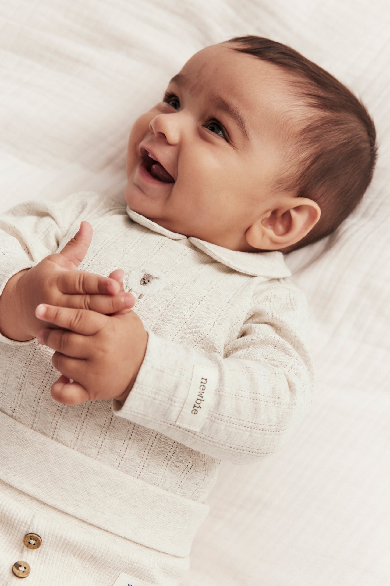 Närbild framifrån på en leende baby i beige melange långärmad pointelle rib body med krage och brodyr.
