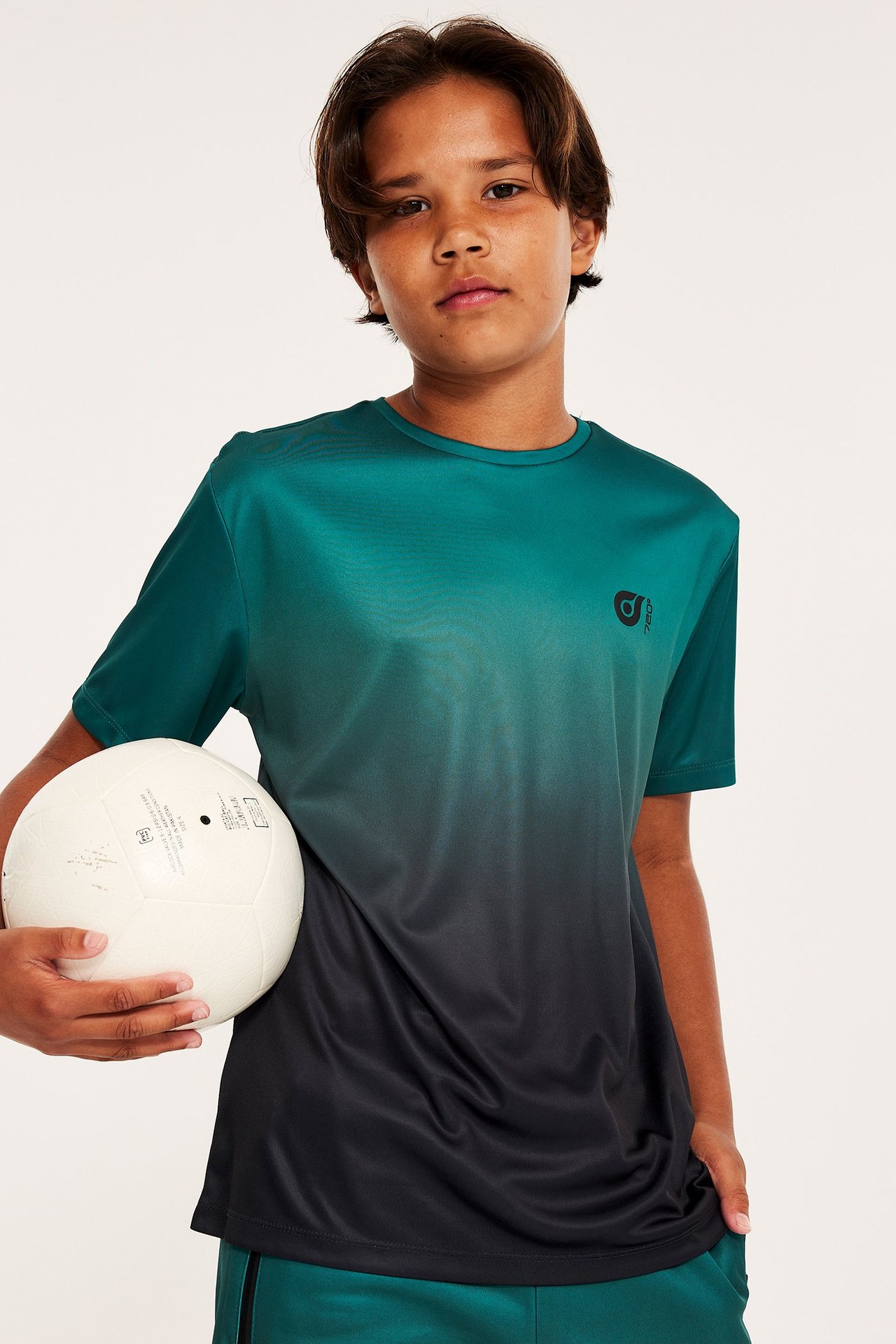 Widok z przodu dziecka w zielonej koszulce sportowej dip-dye, przechodzącej w czerń, trzymającego białą piłkę.