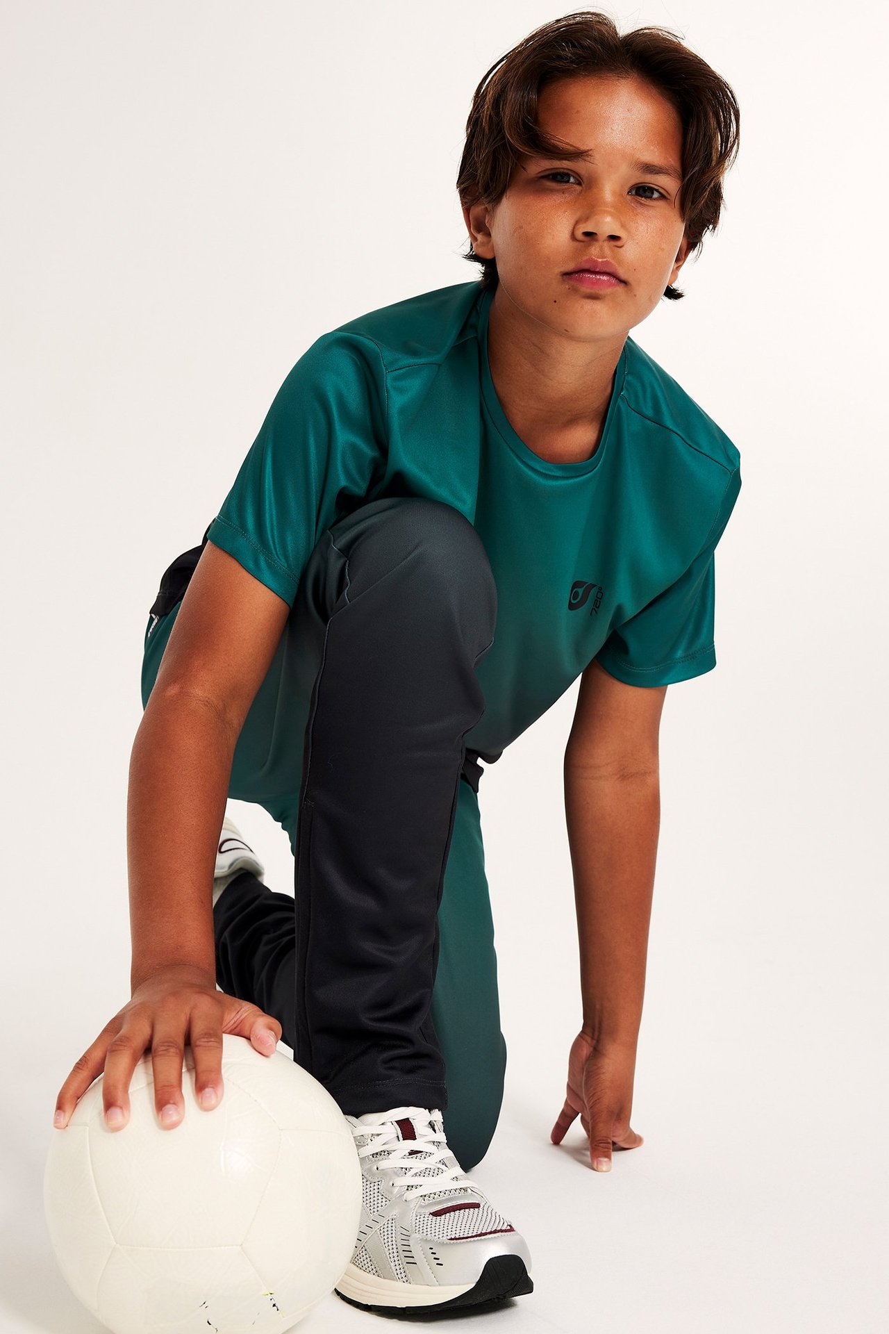 Dziecko w zielonej poliestrowej koszulce sportowej, ciemnoszarych spodniach i sneakersach, z białą piłką.