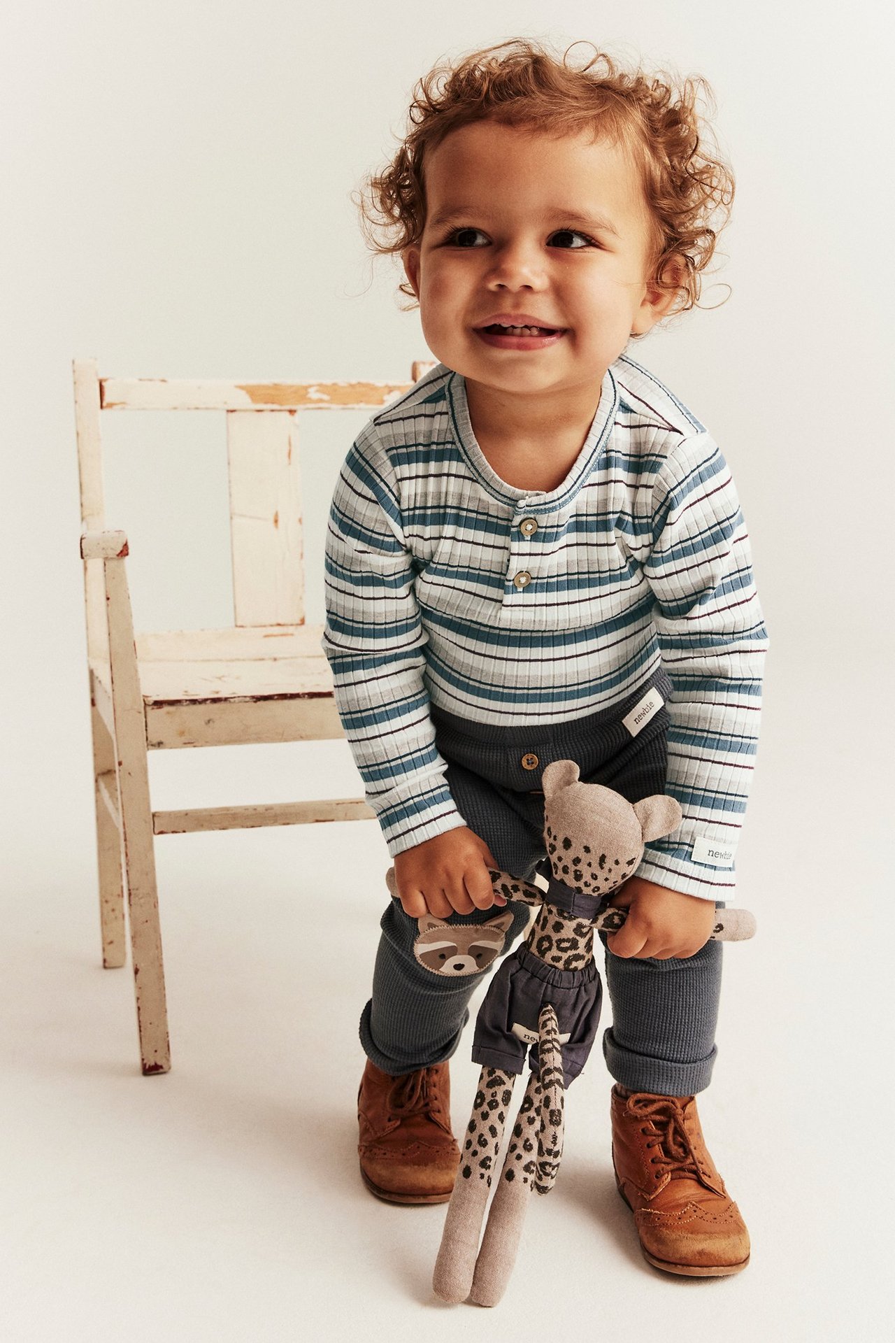 Dziecko w niebiesko-paskowanym prążkowanym body z długim rękawem, guzikami, granatowych legginsach i brązowych butach.