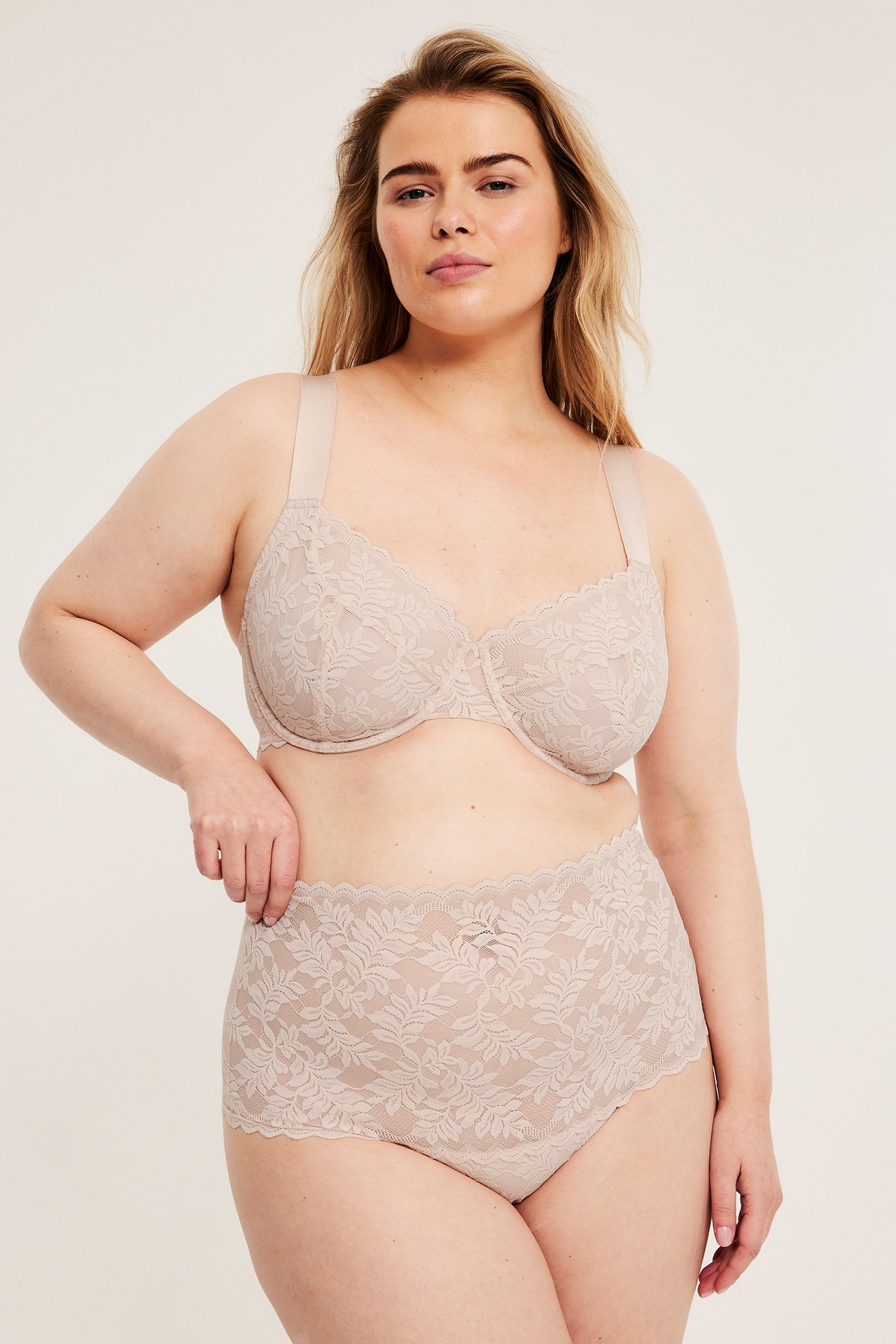 Frontvy av en kvinna i ljusbeige hög midja spetstrosa och en matchande spetsbehå.