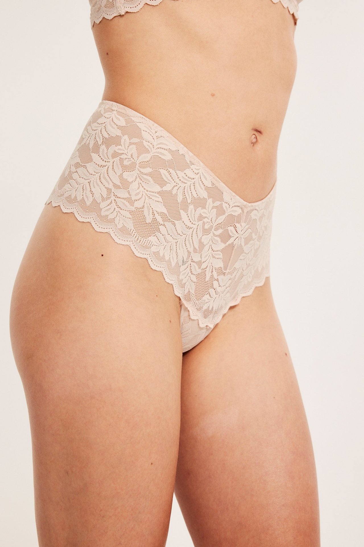 Sidebilde av kvinne i lys rosa V-formet høy midje blondestringtruser og matchende BH.