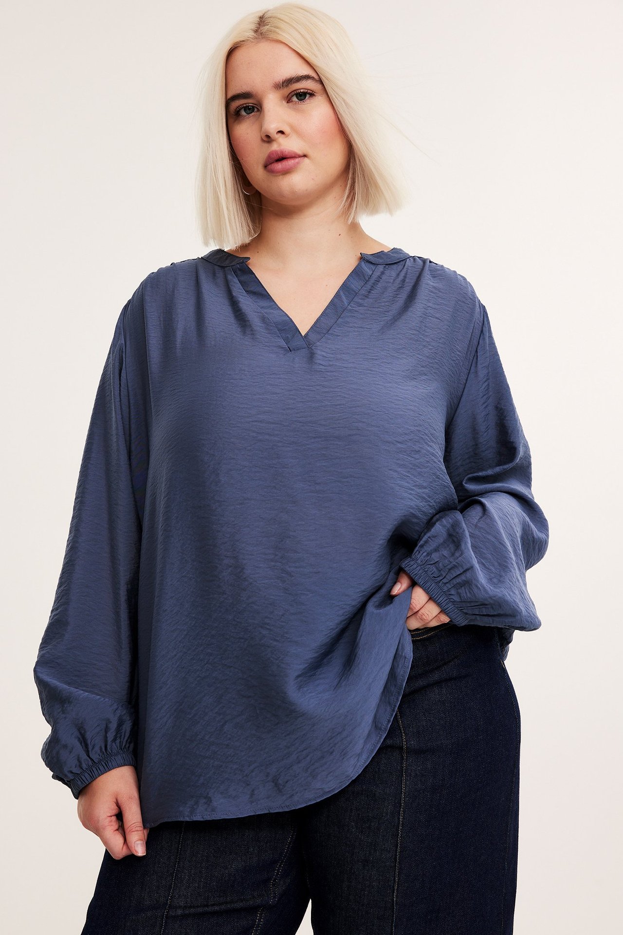 Widok z przodu kobiety w luźnej niebieskiej bluzce z dekoltem w serek, elastycznymi mankietami i ciemnych dżinsach.