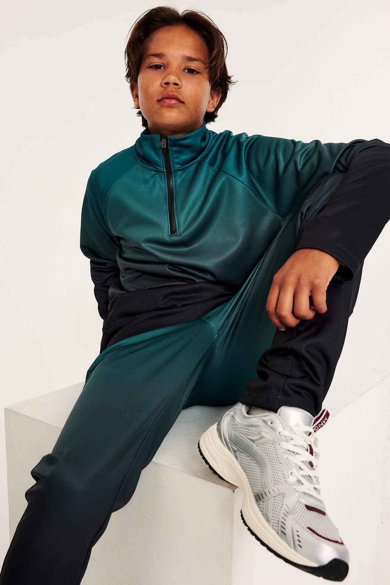 Widok z przodu dziecka w turkusowo-czarnej bluzie treningowej dip-dye z półzamkiem, pasujących spodniach i srebrnych sneakersach.