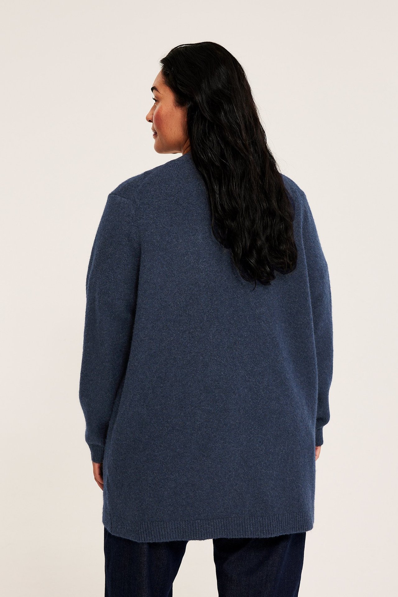 Bakfra av en kvinne i en lang, løs, mørkeblå melert strikket cardigan over mørkeblå jeans.