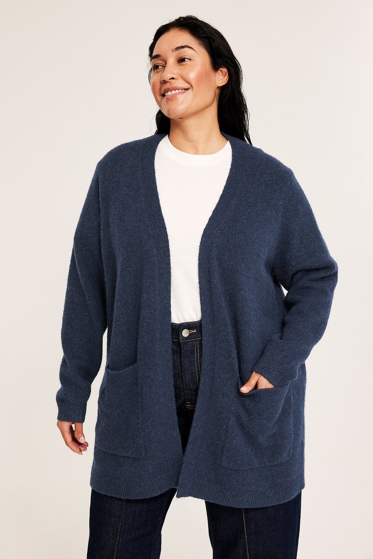 Edestä: Nainen pitkässä, meleeratussa tummansinisessä neuleneuletakissa taskuilla, valkoisen t-paidan ja tummien farkkujen päällä.