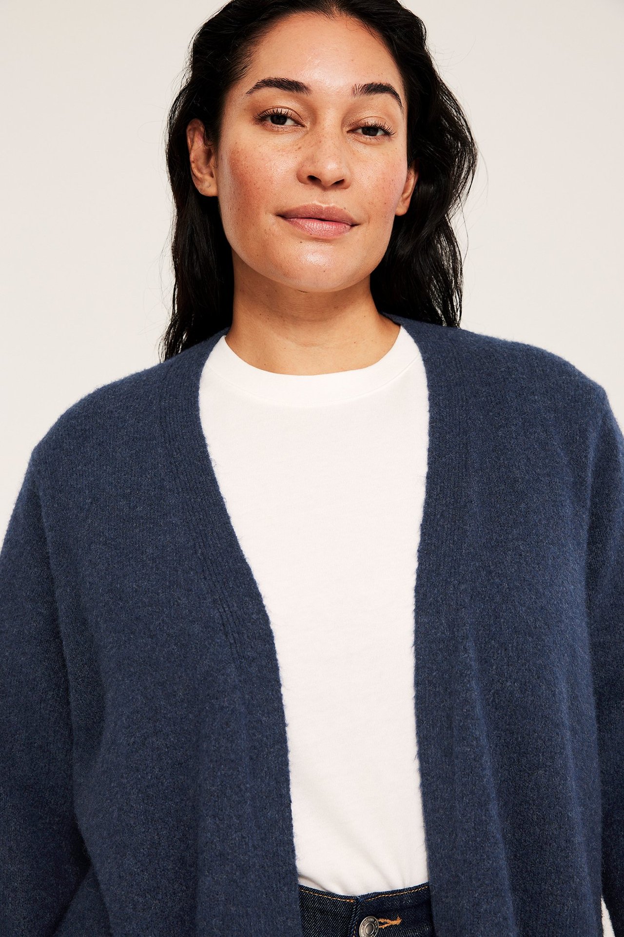 Lähikuva edestä naisesta, jolla on avoin tummansininen meleerattu neulejakku valkoisen t-paidan päällä.