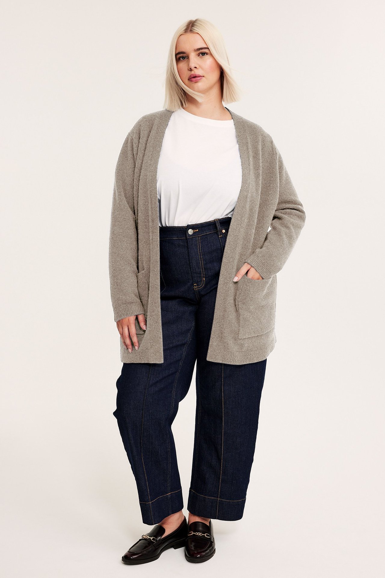 Kvinna bär en lång beige melerad stickad kofta med fickor, vit t-shirt, mörkblå jeans, svarta loafers.