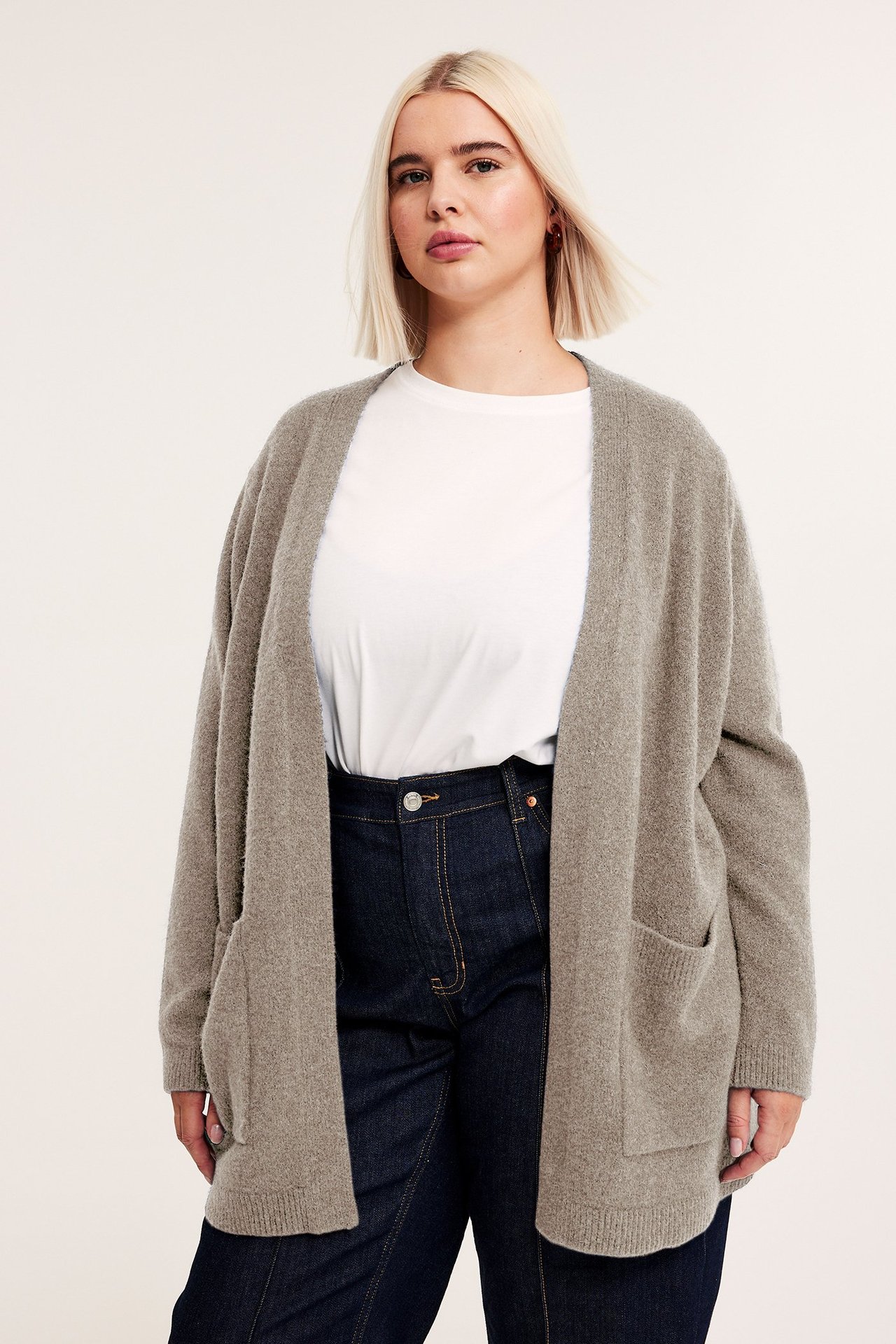 Frontvy: Kvinna i lång, öppen, melerad beige stickad kofta med fickor, vit t-shirt, mörka jeans.