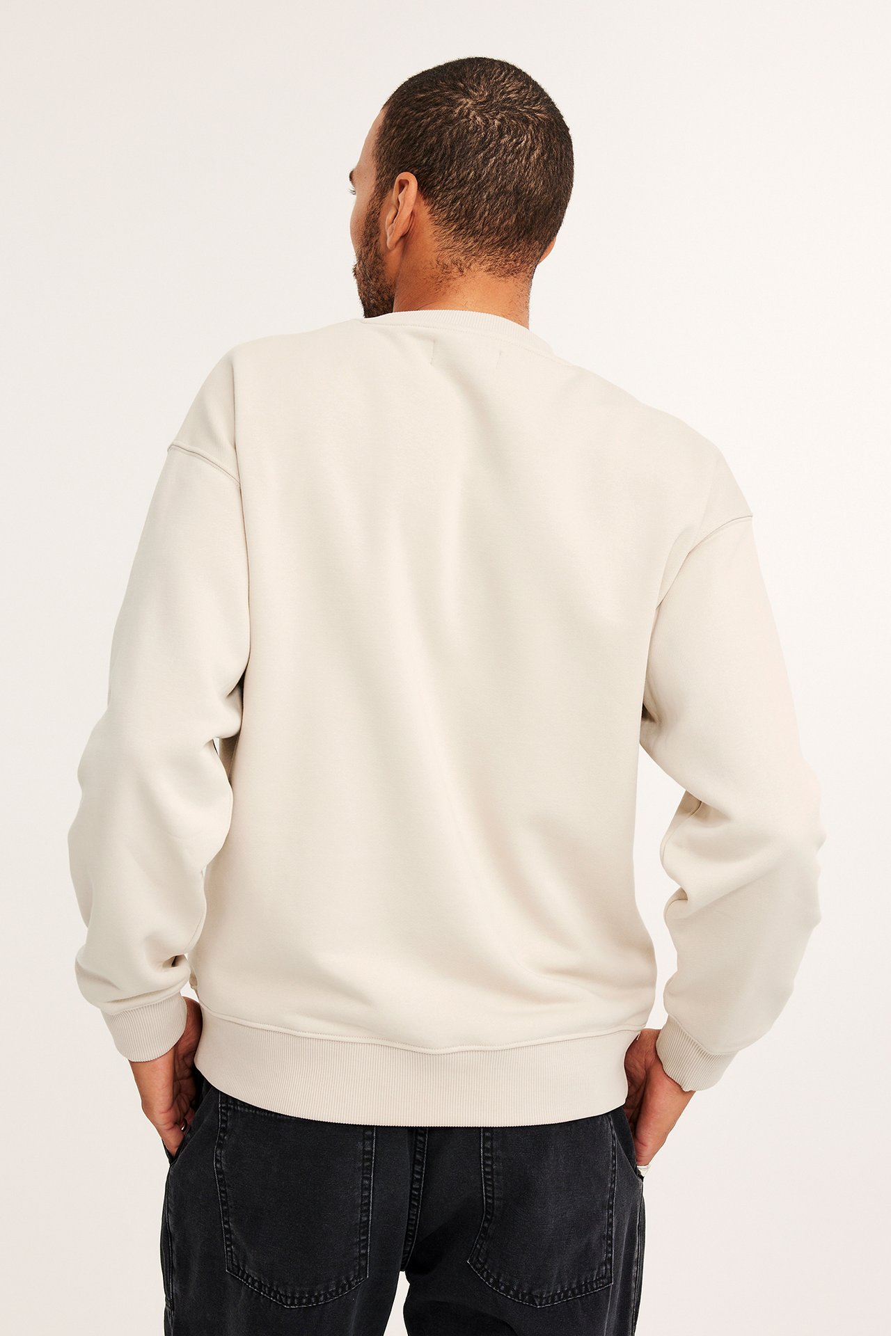 Bakfra, mann i lys beige oversized sweatshirt med senkede skuldre og mørke jeans.