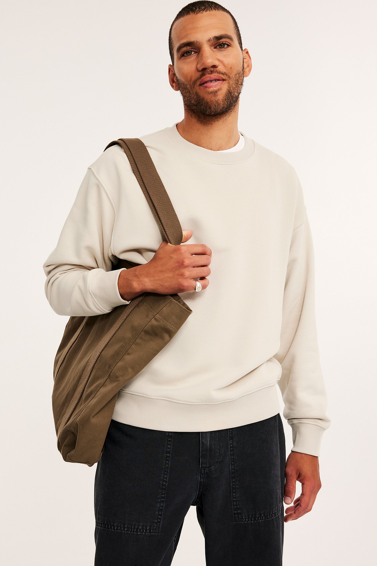 Frontbilde av en mann i en oversized lys beige sweatshirt, mørkegrå bukser, holdende en khaki totebag.