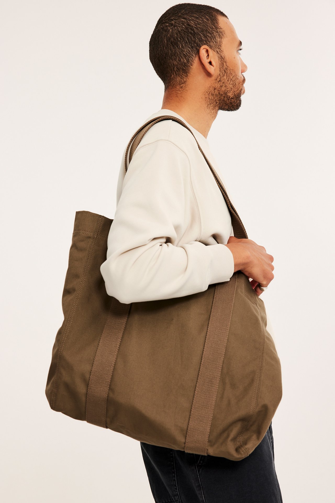 Sideprofil av en mann i en lys beige oversized sweatshirt, bærende en stor brun stoffveske.