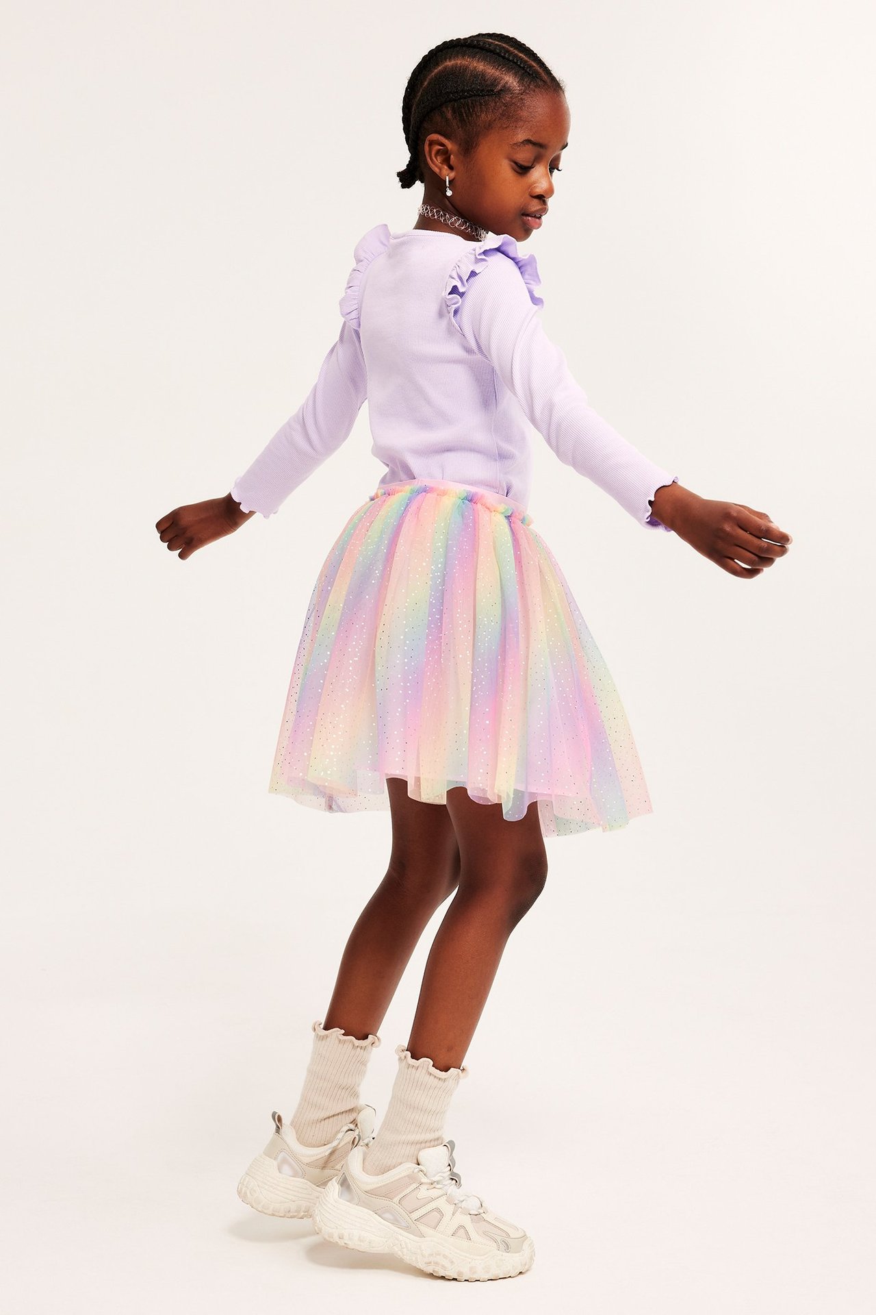 Sivukuva lapsesta sateenkaarenvärisessä glittertyllihameessa, vaaleanliilassa röyhelötopissa, beigenvärisissä sukissa ja lenkkareissa.