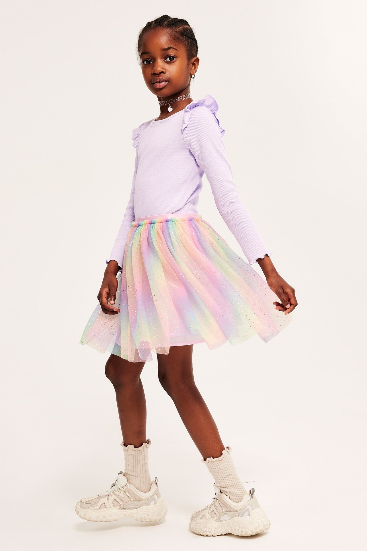 Widok z przodu: Dziecko w tęczowej tiulowej spódnicy z brokatem, jasnofioletowej bluzce z falbankami i jasnobeżowych sneakersach.