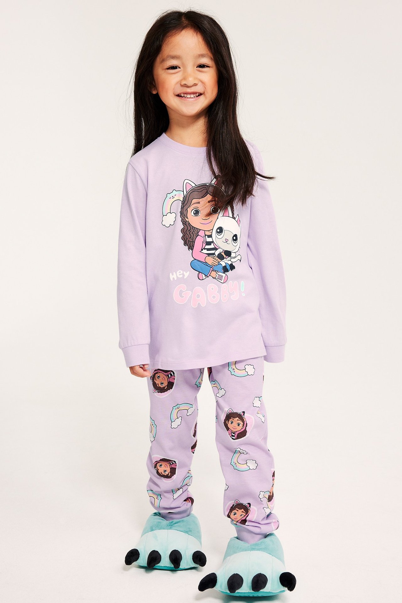 Lapsi tummanliilaloisissa pyjamissa Gabby's dollhouse -kuviolla, housut rainboilla ja pilvillä, tossut.