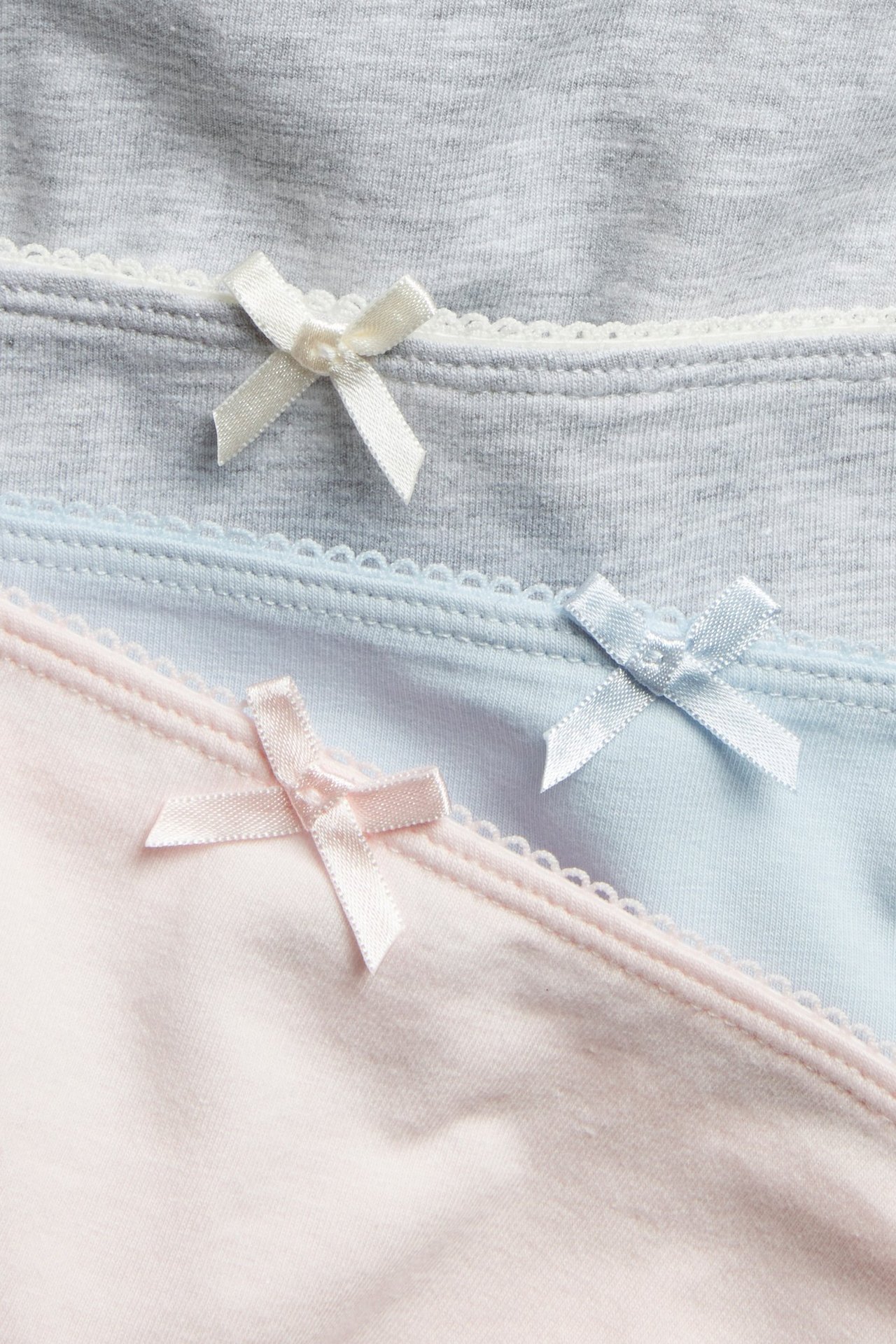 Zbliżenie na trzy jednokolorowe majtki w jasnym różu, błękicie i szarości, z brzegami picot i kokardkami.