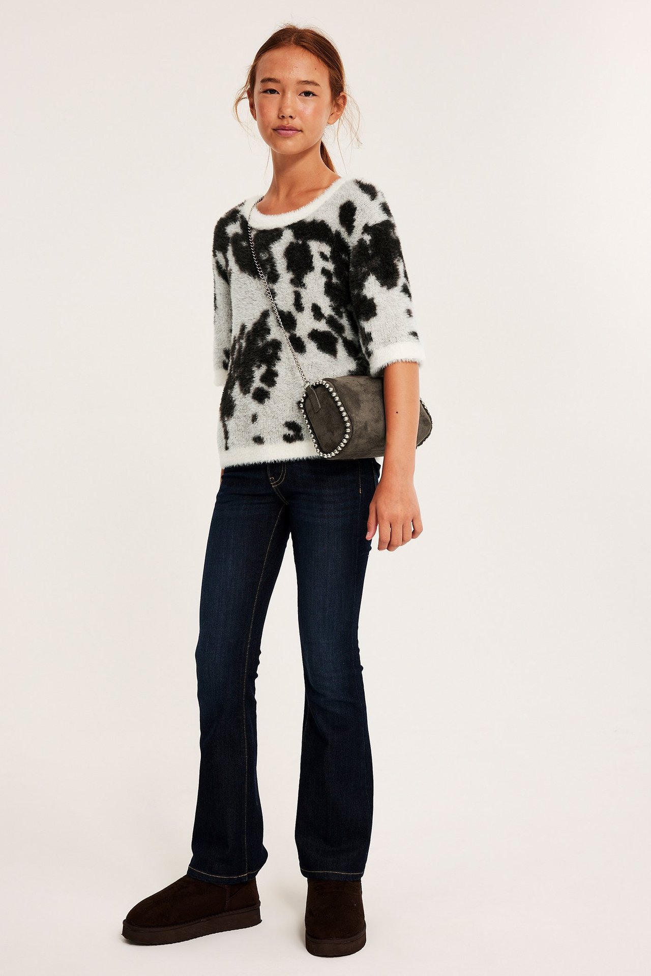 Frontvy: Barn i off-white, svart jacquard fluffig kortärmad tröja, blå utsvängda jeans, bruna stövlar, grå väska.