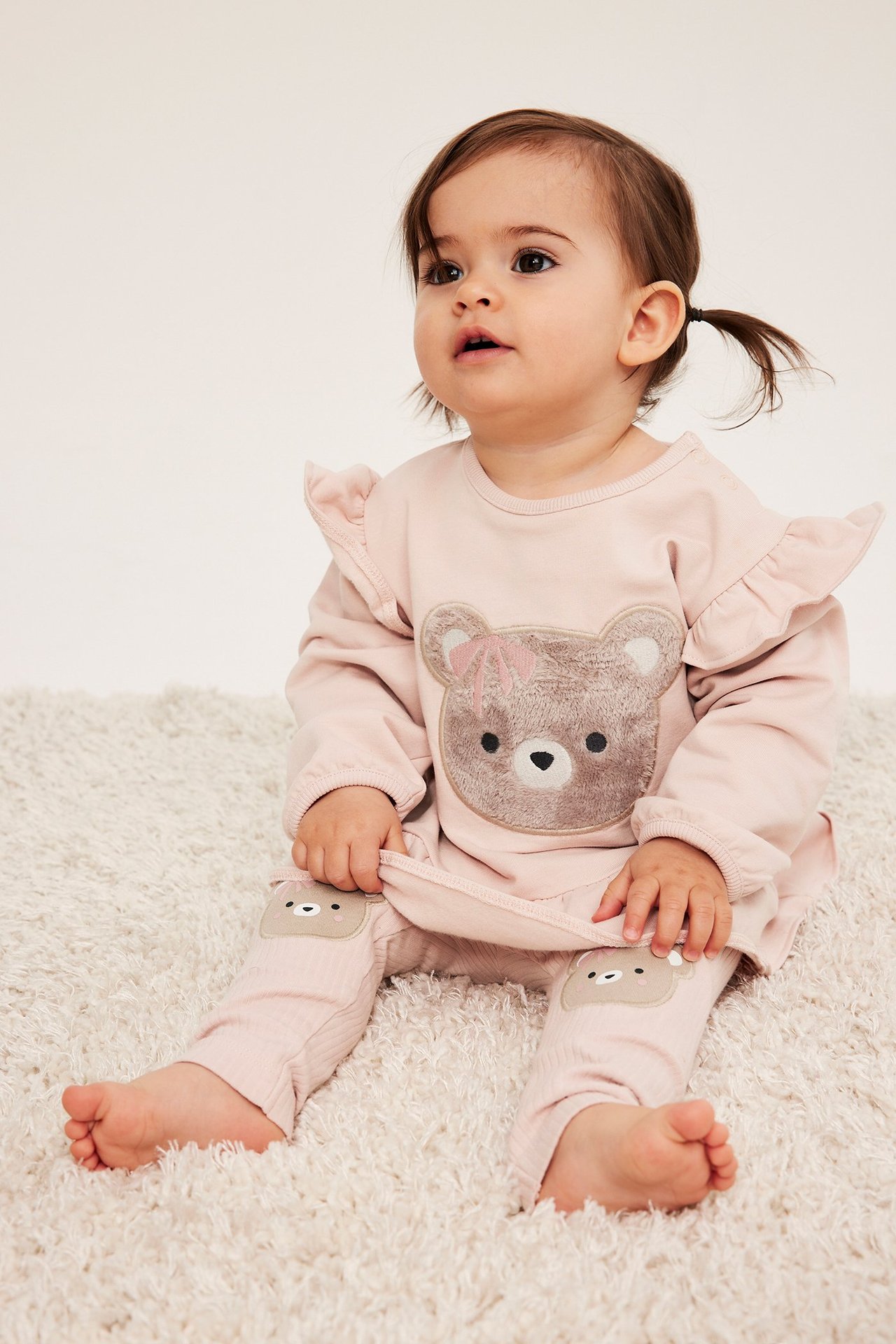 Edestä, vauva vaaleanpunaisissa ribatuissa leggingseissä karhuapplikaatioilla ja pitkähihaisessa topissa.