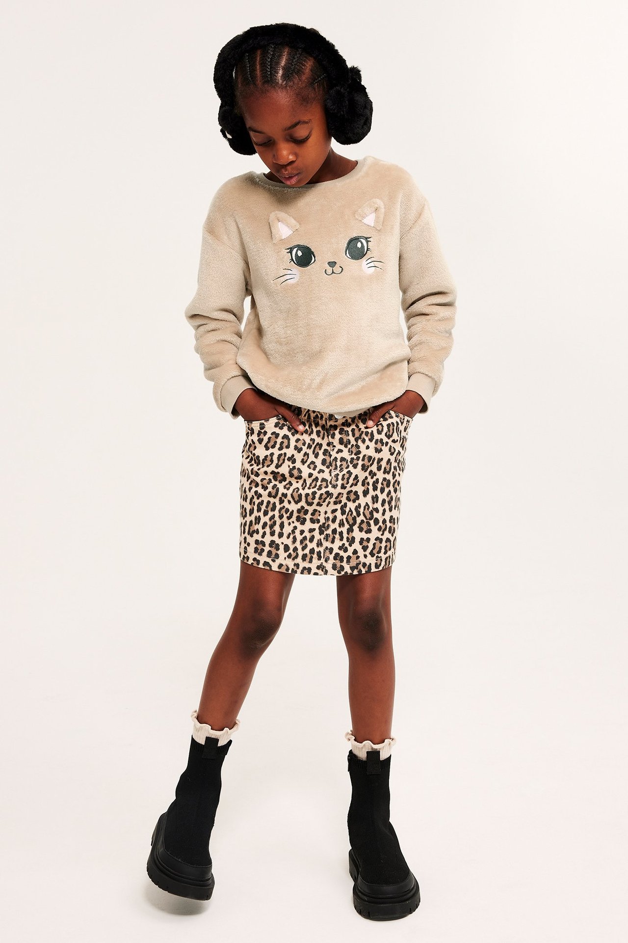 Frontvy av ett barn i beige fluffig tröja med kattapplikation, leopardmönstrad kjol, svarta kängor och svarta hörselkåpor.