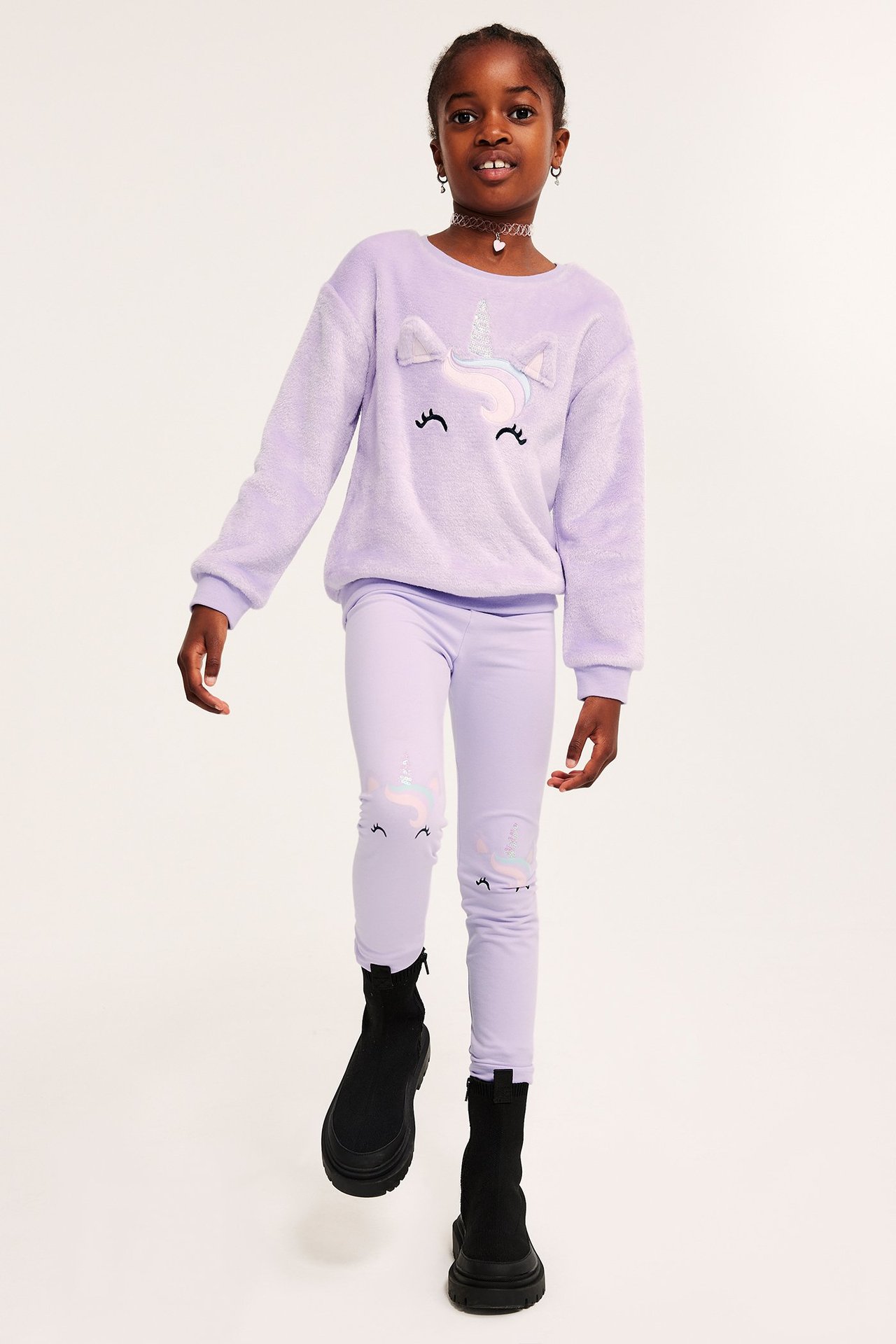 Forfra: barn i lys lilla fluffy enhjørning-sweatshirt, matchende leggings og svarte støvler.