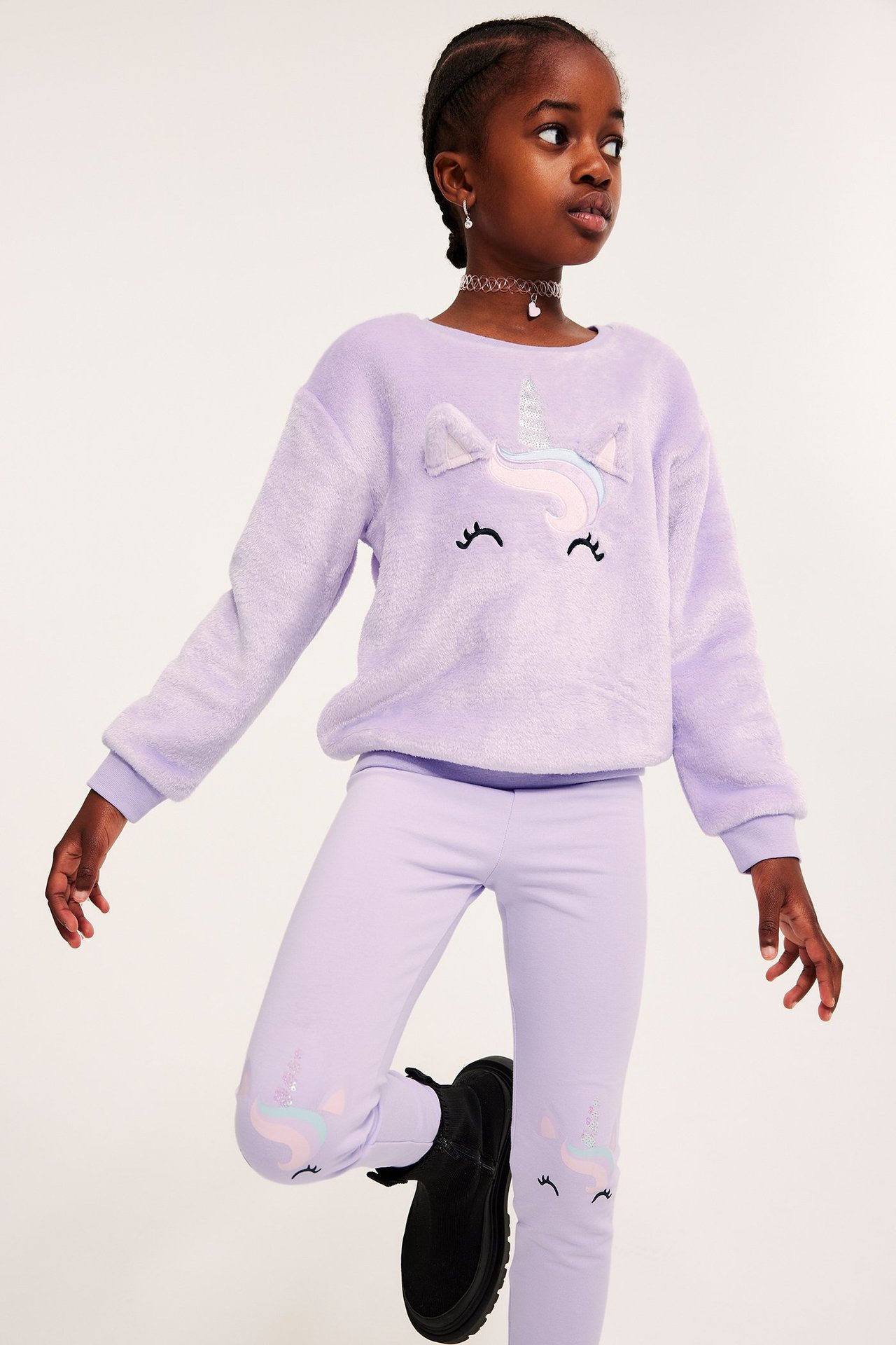 Widok z przodu: dziecko w jasnofioletowej puszystej bluzie z jednorożcem, legginsach i czarnych butach.