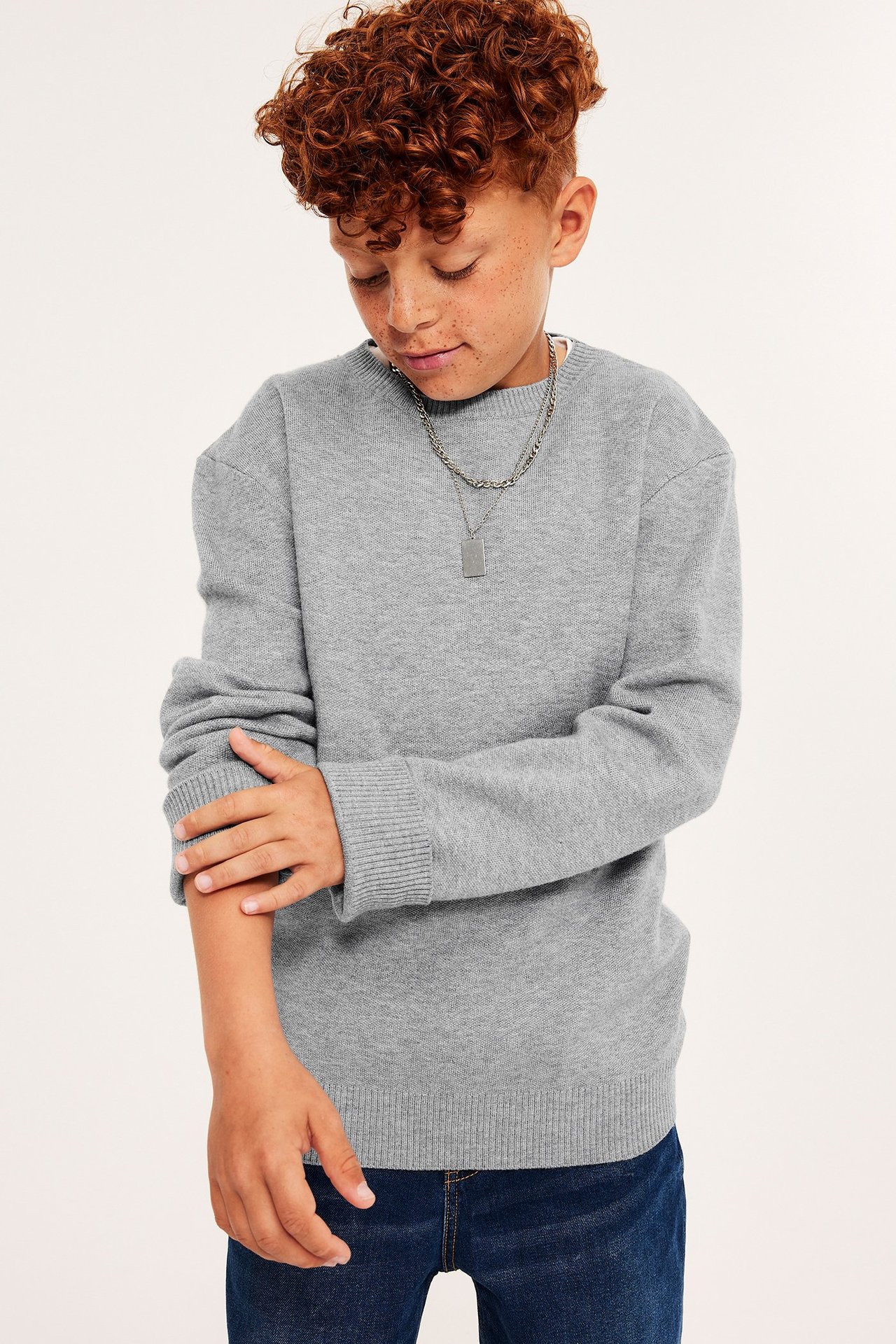 Frontvy av ett barn i gråmelerad stickad rundhalsad tröja, mörkblå jeans och silverfärgat halsband.