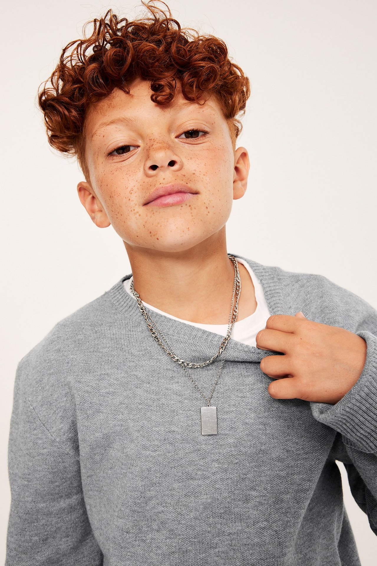 Framifrån: barn i mörkgrå melerad stickad tröja, vit t-shirt och silverhalsband.