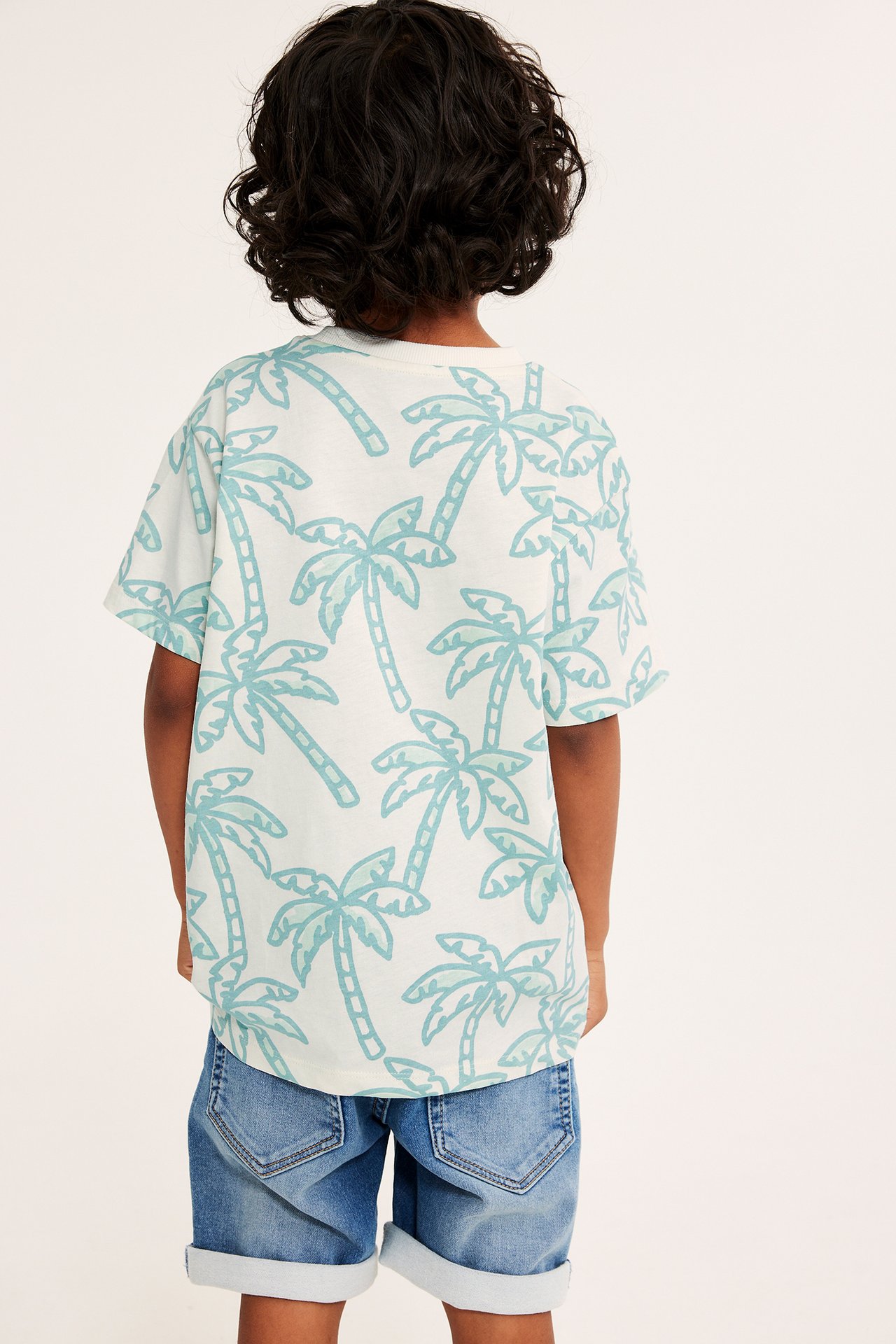 Widok z tyłu: Dziecko w kremowym, oversize'owym bawełnianym T-shircie z jasnoniebieskim wzorem palm i niebieskich szortach.