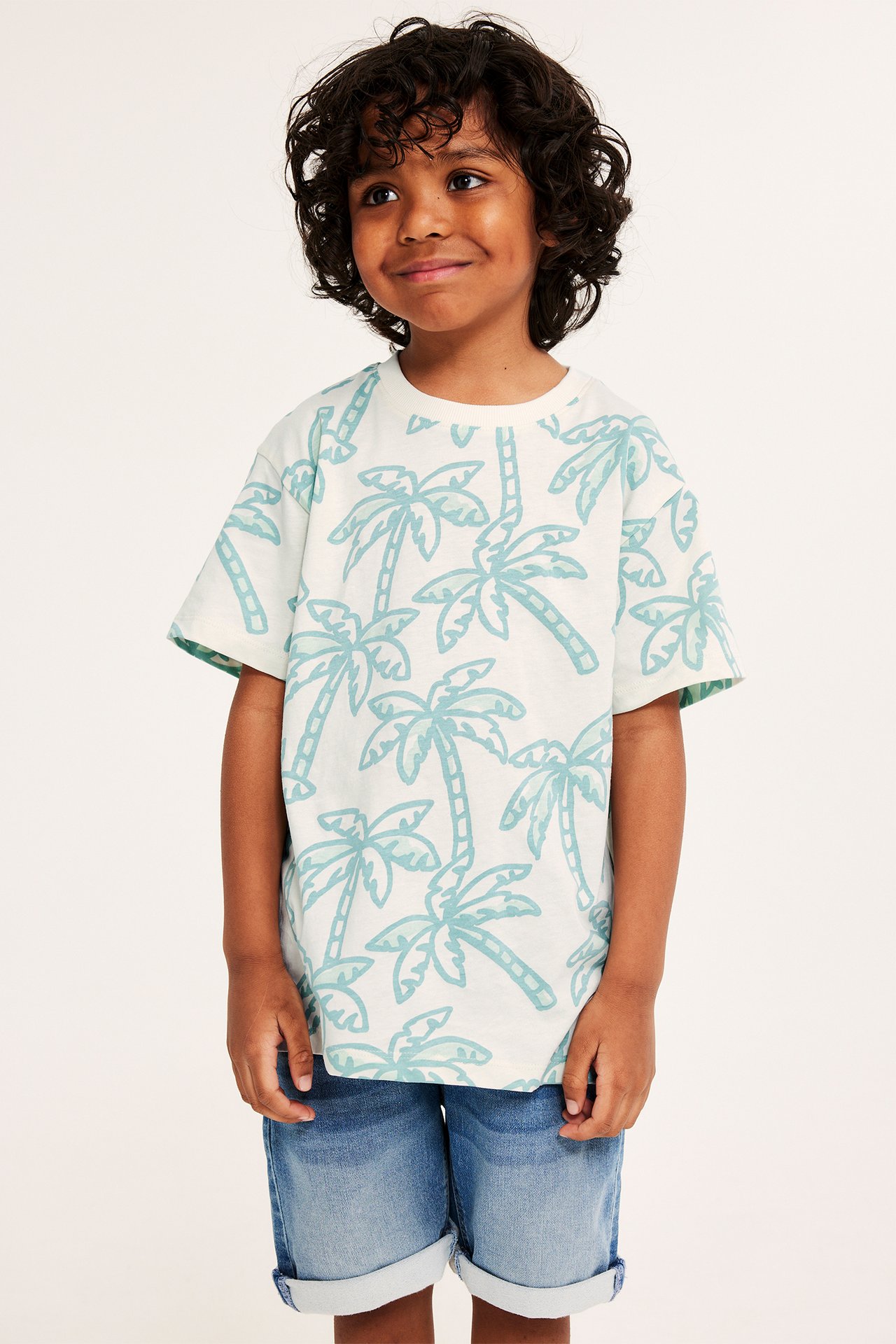 Widok z przodu: Dziecko w kremowym bawełnianym T-shircie oversize z jasnoniebieskim wzorem palm i niebieskich szortach.