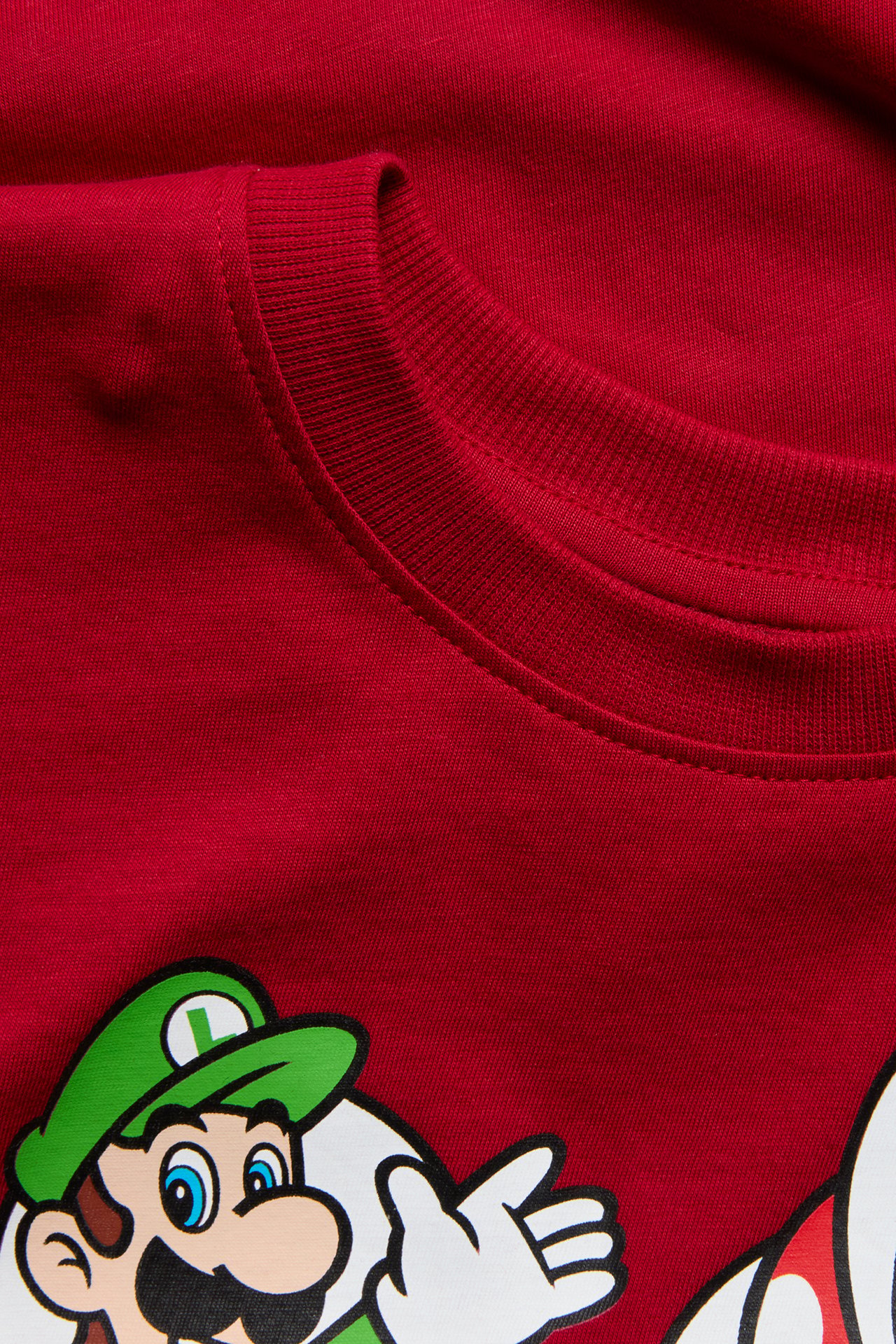 Lähikuva lasten punaisesta puuvilla-T-paidasta, jossa on ribattu pääntie ja Super Mario -painatus.