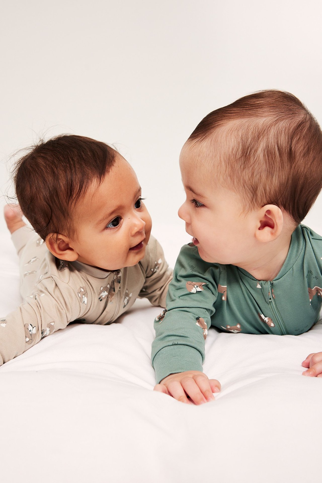 Kaksi vauvaa kuviollisissa potkupuvuissa: beige ja vihreä kettupainatuksella, kaksisuuntaisilla vetoketjuilla.