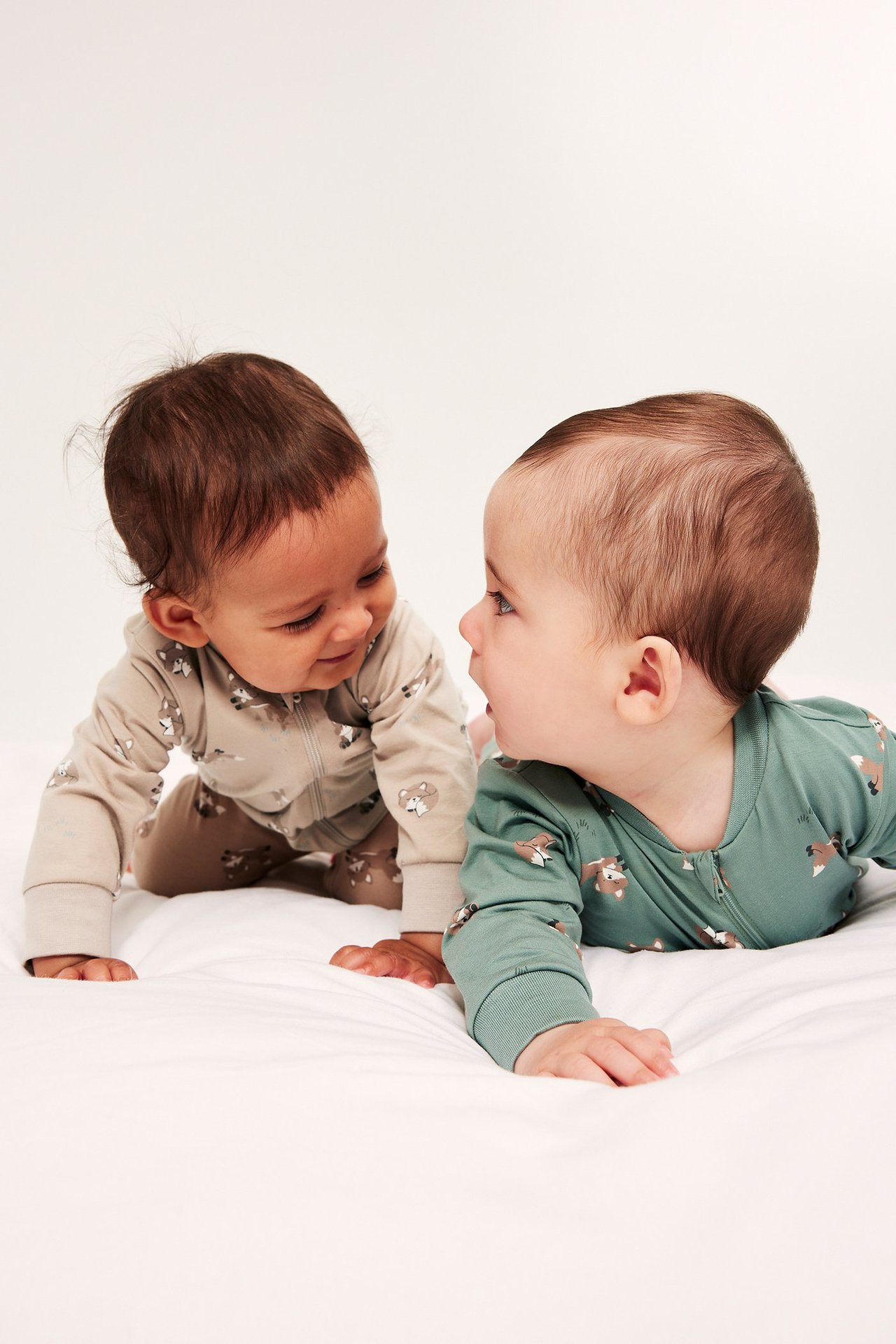 Edestä: kaksi vauvaa, toinen beigessä kuviollisessa potkupuvussa kaksisuuntaisella vetoketjulla, toinen vihreässä.