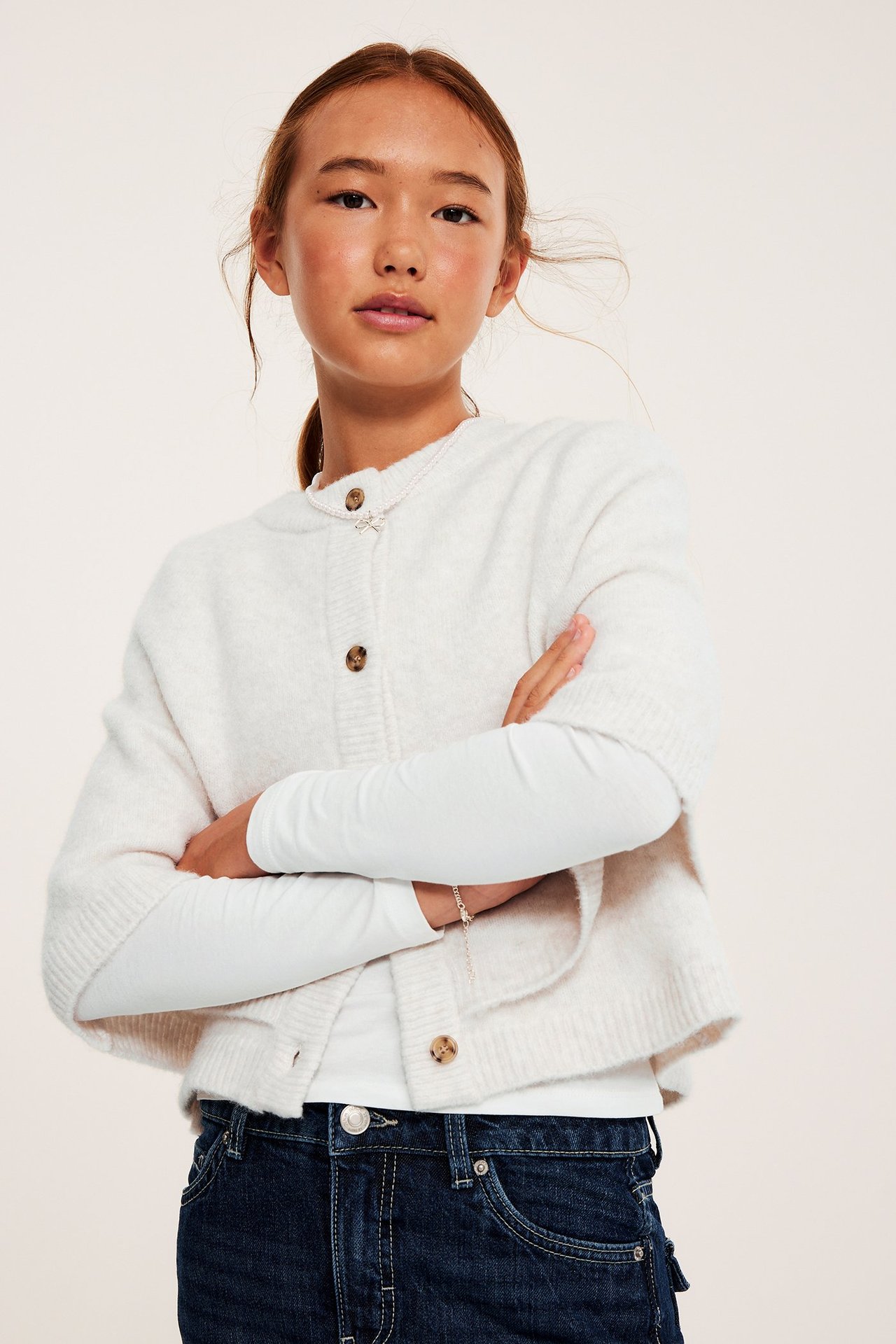 Widok z przodu: Dziecko w melanżowym białym ponczo z dzianiny, z brązowymi guzikami, na białym topie i dżinsach.
