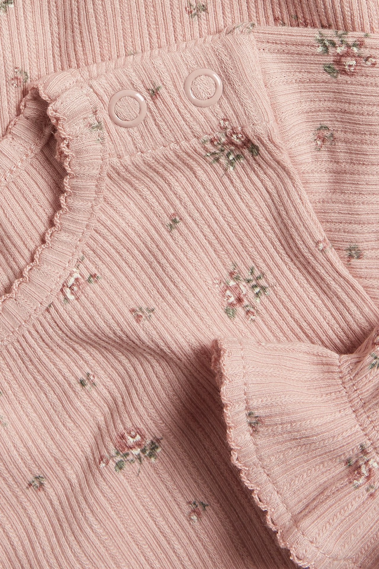 Lähikuva vaaleanpunaisesta ribbineulotusta vauvan body-paidasta, jossa kukkakuvio, picot-reuna ja nepparit.
