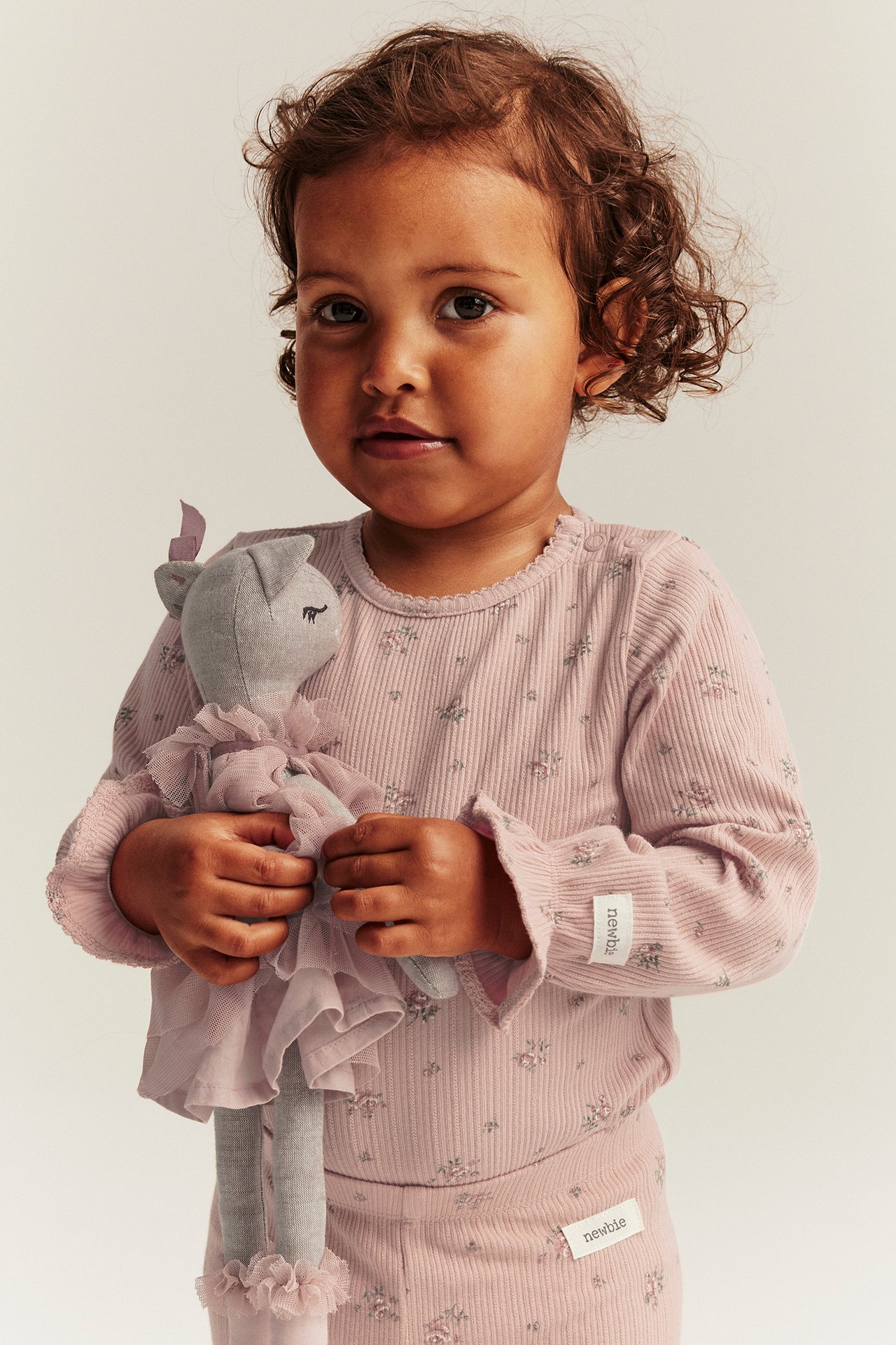Frontbilde av en babyjente i rosa ribbet blomstret langermet body og matchende bukser, holder en grå dukke.