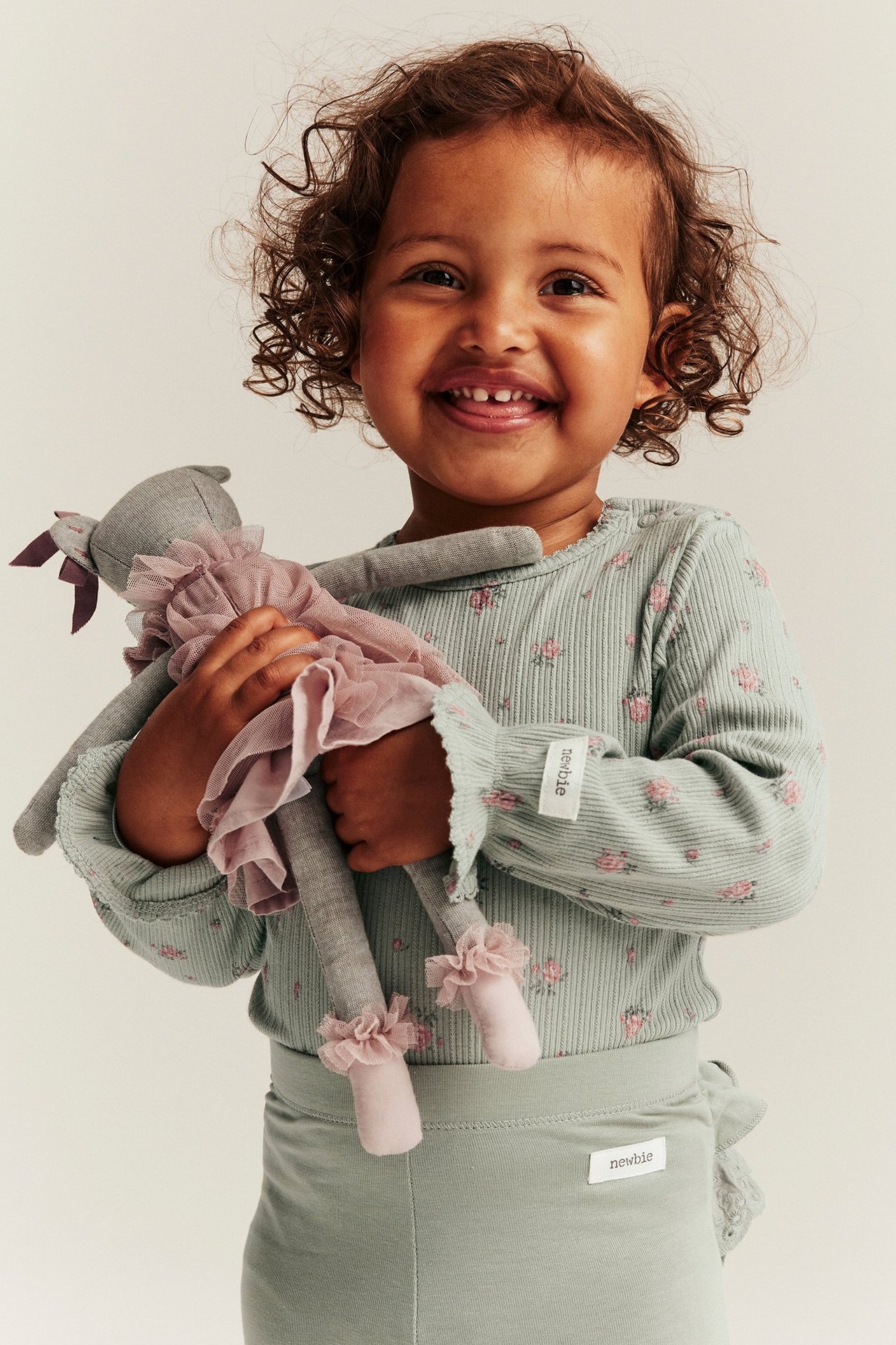 Lyhyet kiharat ruskeat hiukset omaava vauva. Yllään vihreä kukkakuvioinen body ja vihreät leggingsit. Lapsi pitelee pehmolelua ja nauraa.